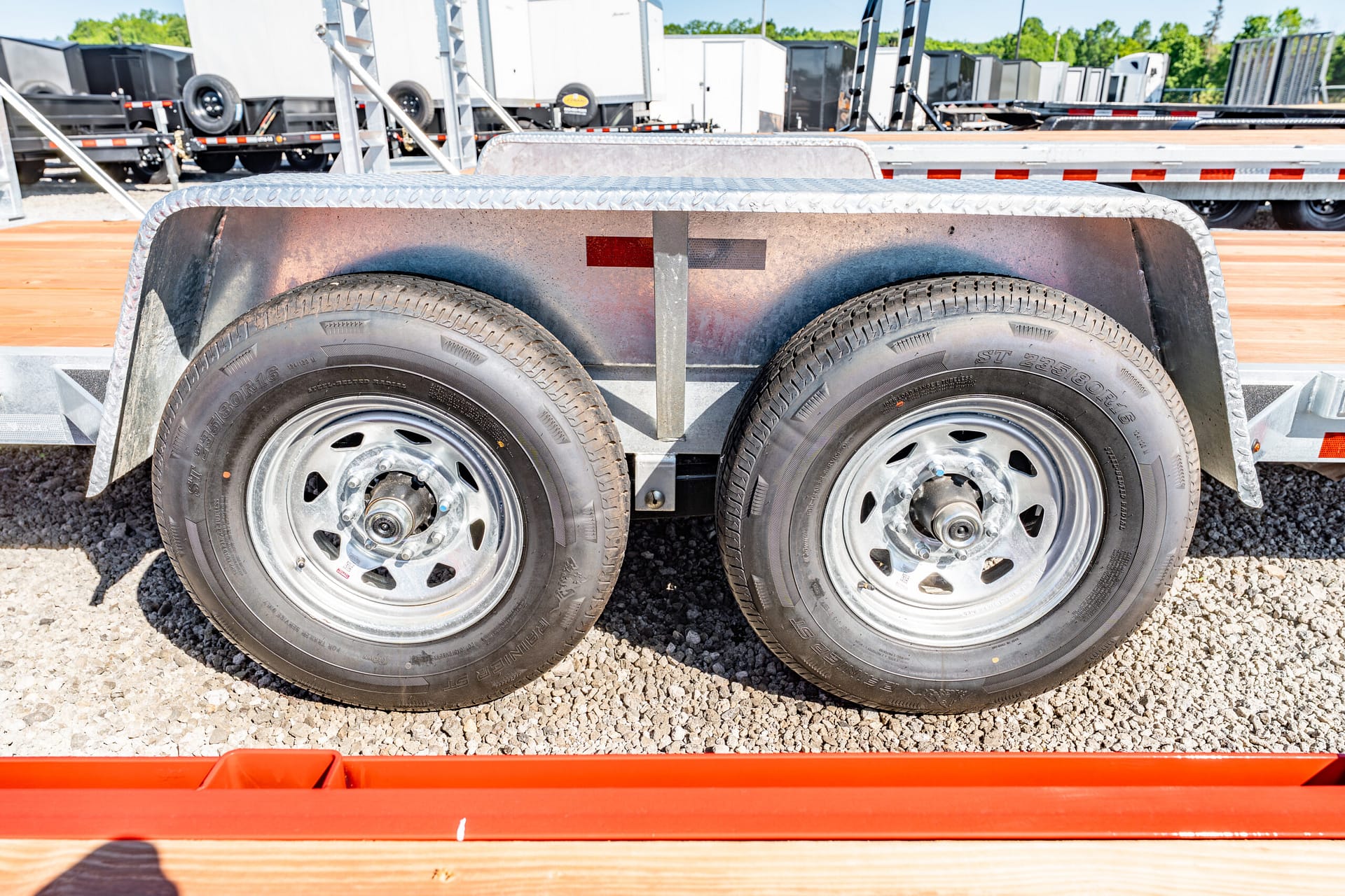 Dual trailer wheels with silver rims on a metal utility trailer, parked on gravel.