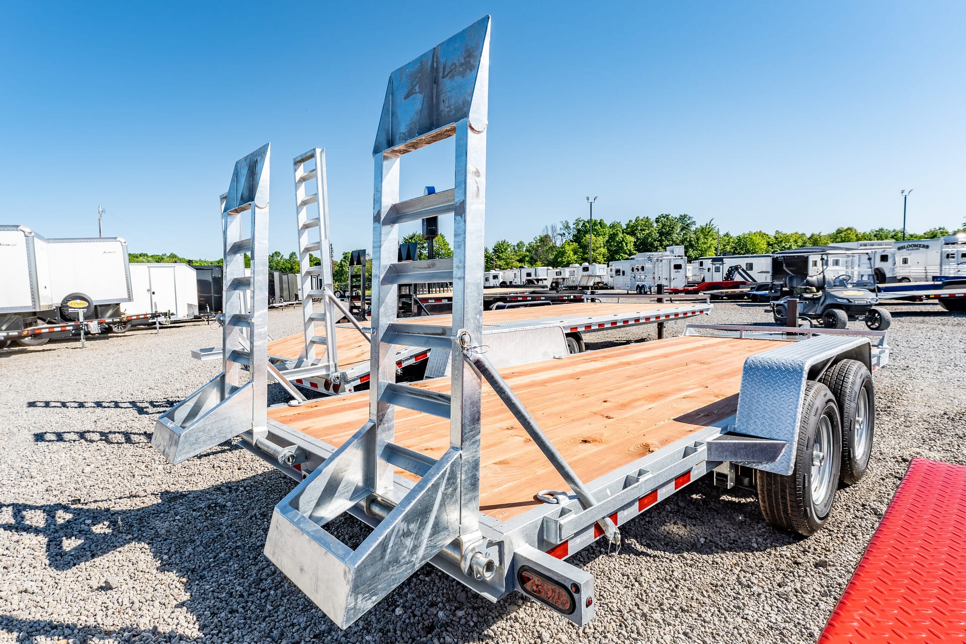 Flatbed trailer with metal ramps and wooden deck in a gravel lot, clear blue sky above.