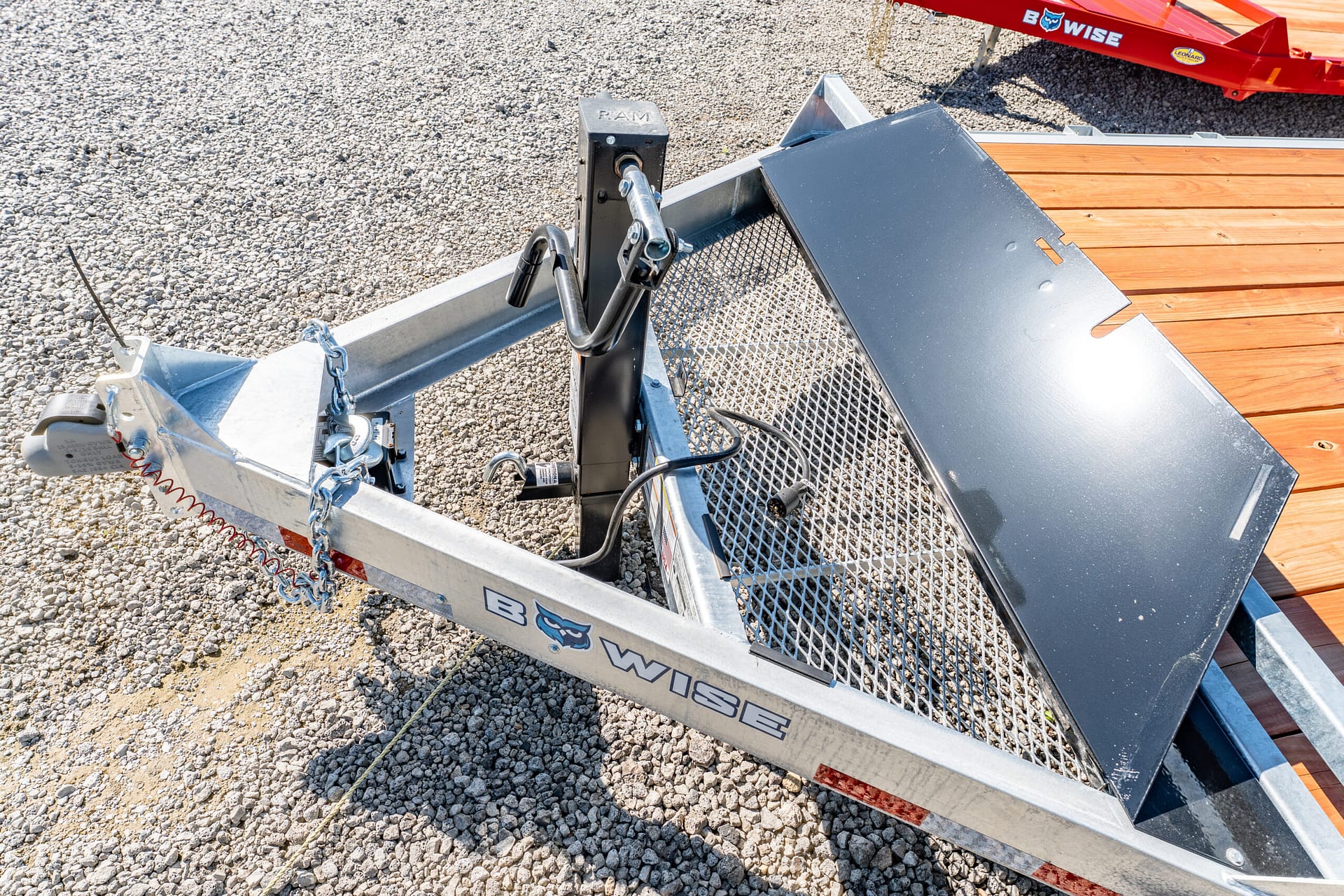 Close-up of a metal utility trailer ramp and jack on a gravel surface with wooden deck planks nearby.