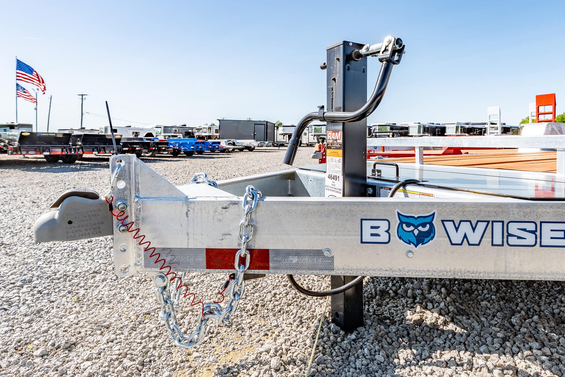Close-up of a metal trailer hitch with safety chains on a gravel lot; trailers and American flags in the background.