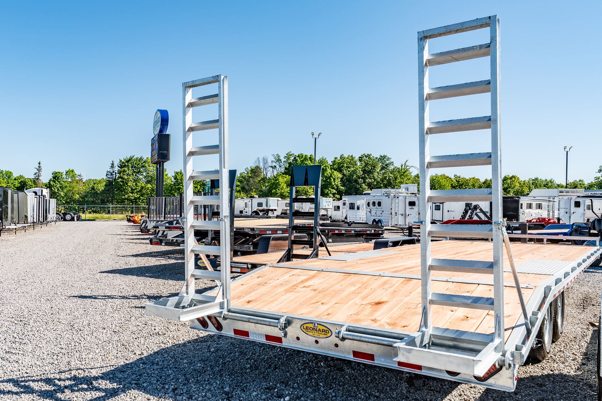 Outdoor equipment yard with aluminum flatbed trailers and wooden decks, under a clear blue sky; several trailers and RVs in the background.