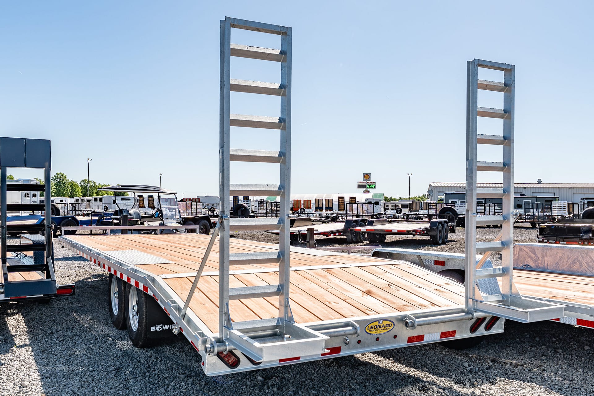 Flatbed trailer with wooden deck in a truck yard, surrounded by equipment and tall metal frames against a clear blue sky.