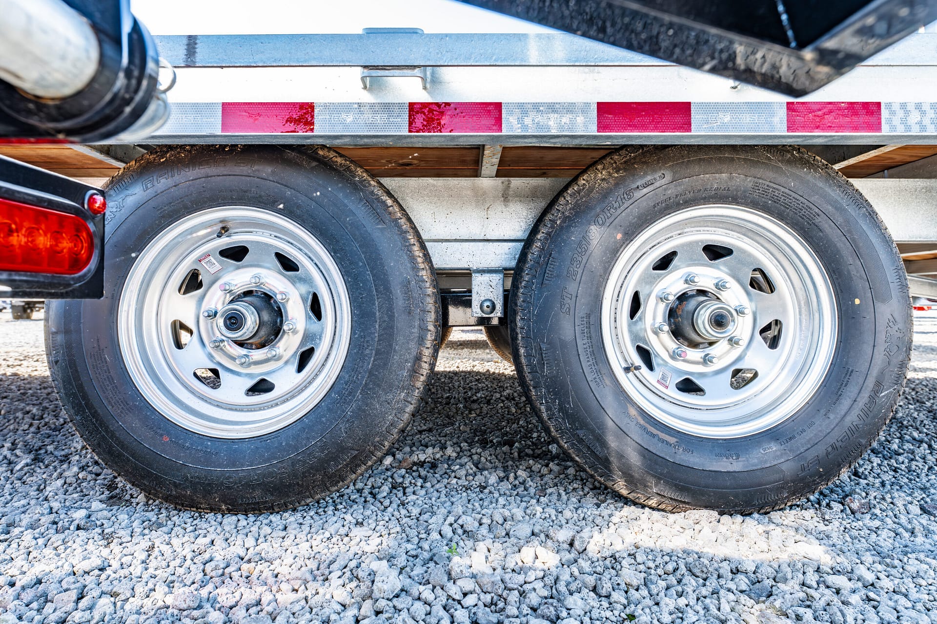 Two tandem trailer wheels with metal rims under a trailer frame, resting on gravel with a red tail light visible on the left.