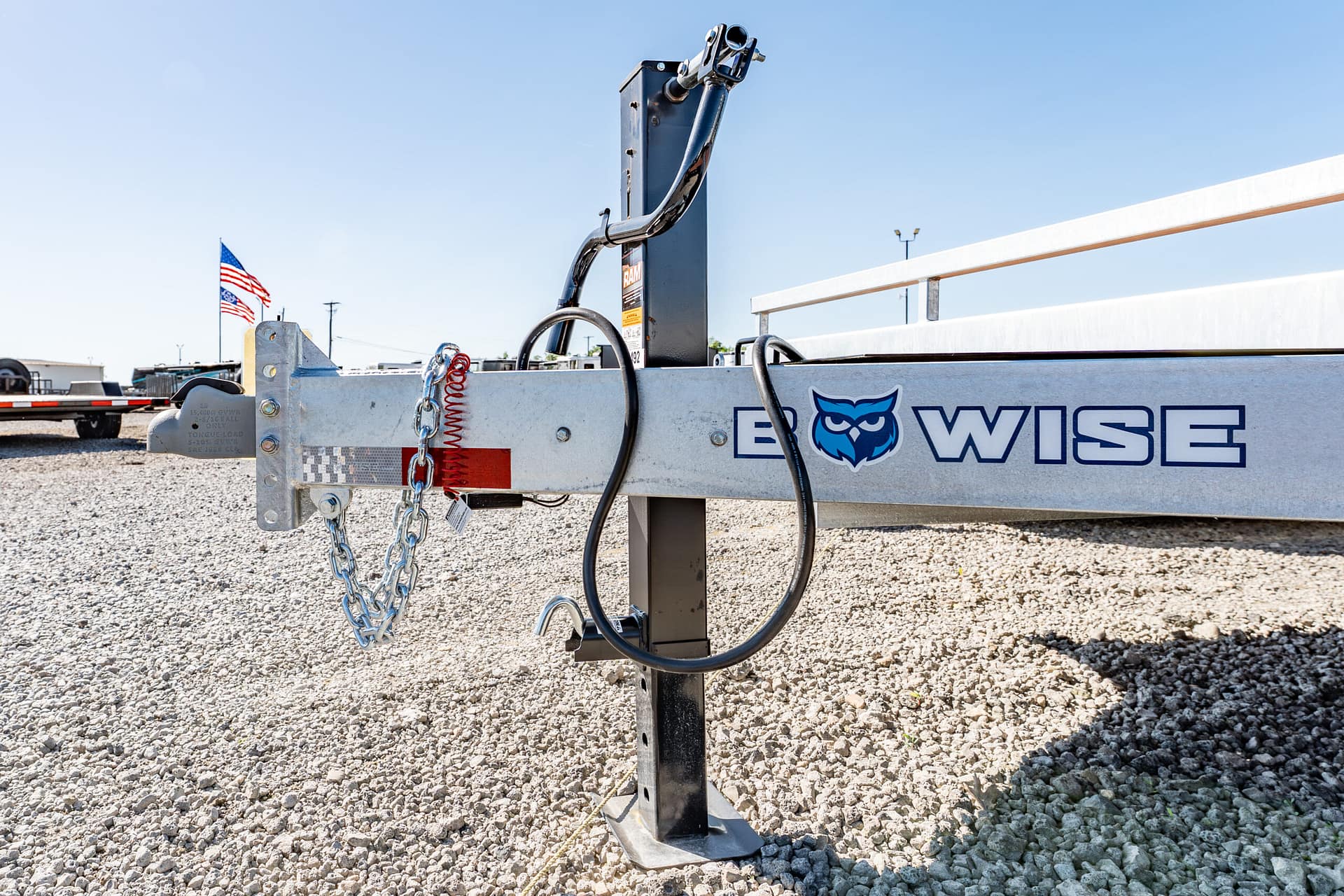 Trailer hitch equipment with chains and metal beam on gravel, American flags in the background and a blue sky, branding 'WISE' on the trailer rail.
