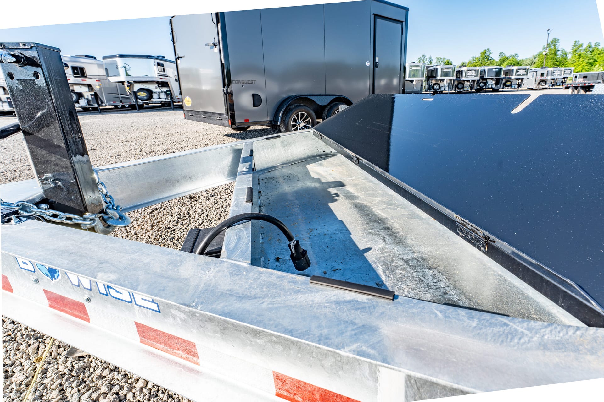 Close-up of a metal loading ramp on a flatbed trailer with safety chains, parked among other trailers in a gravel lot.