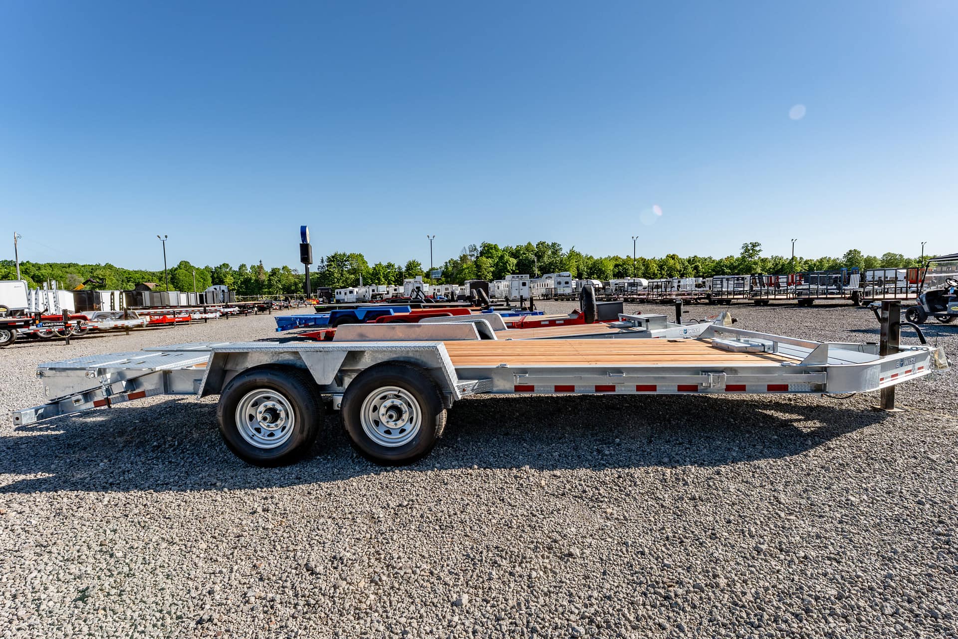 Row of metal trailer beds parked on a gravel lot under a clear blue sky, with trees in the background and various trailers stretching into the distance.
