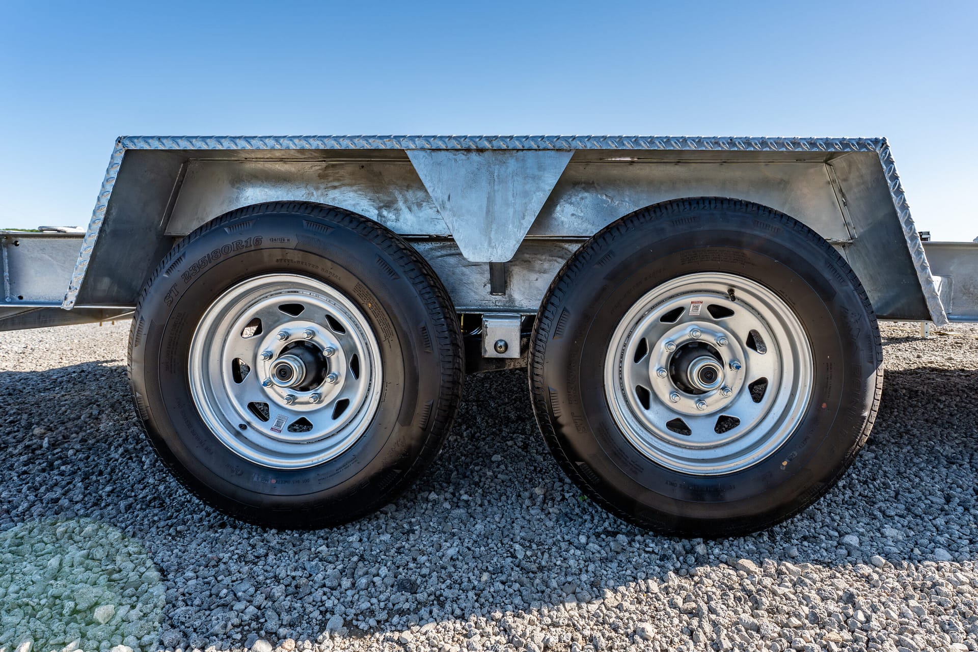 Two large trailer wheels with a diamond-plate metal fender, parked on gravel under a clear blue sky.