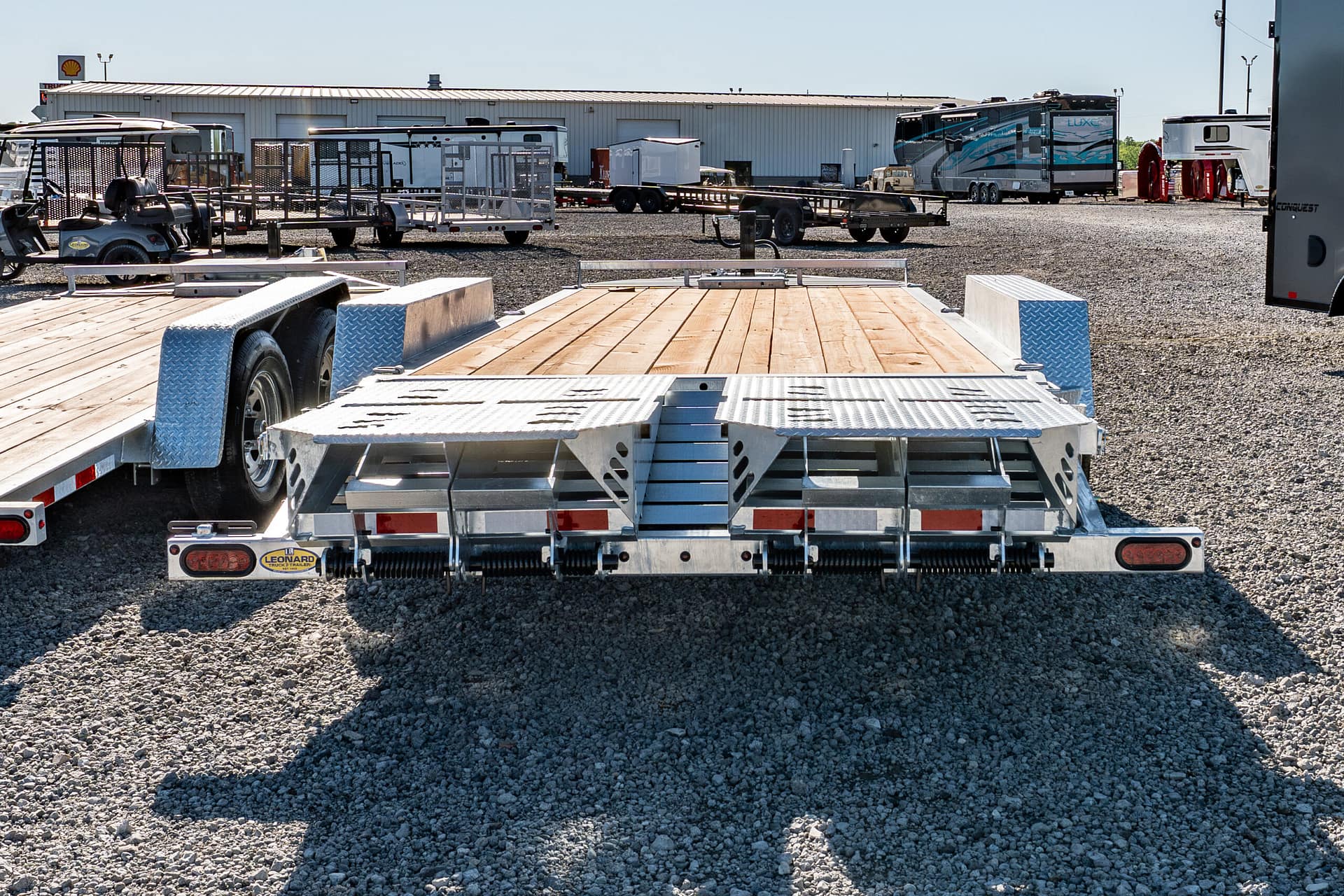 Wide view of a gravel lot with multiple trailers and campers parked near a large metal building in the background.|Note: foreground shows a flatbed trailer with diamond-plate edges and wooden planks