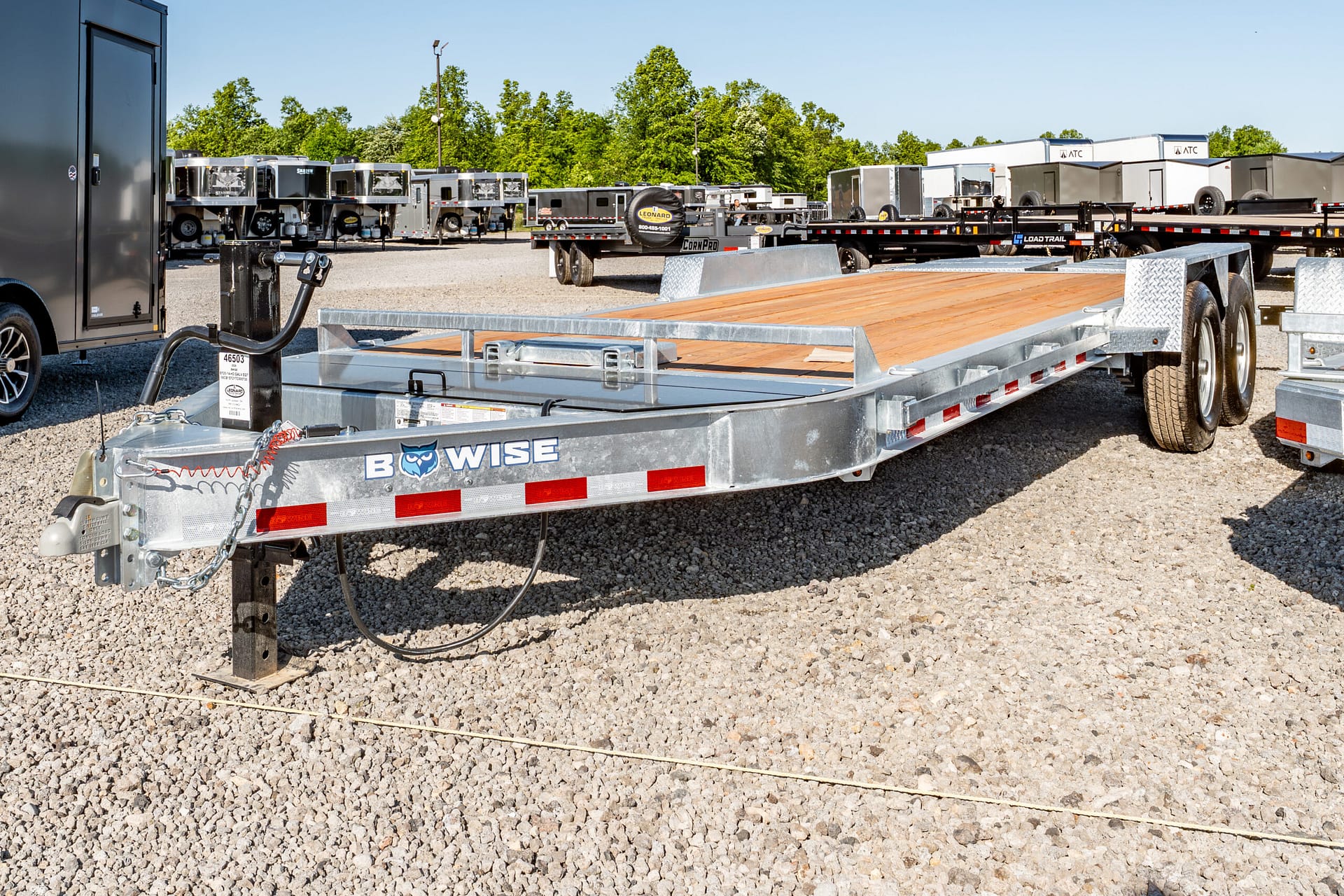 Flatbed trailer with a wooden deck in a gravel lot, surrounded by other trailers and equipment under a clear blue sky on a trailer lot.