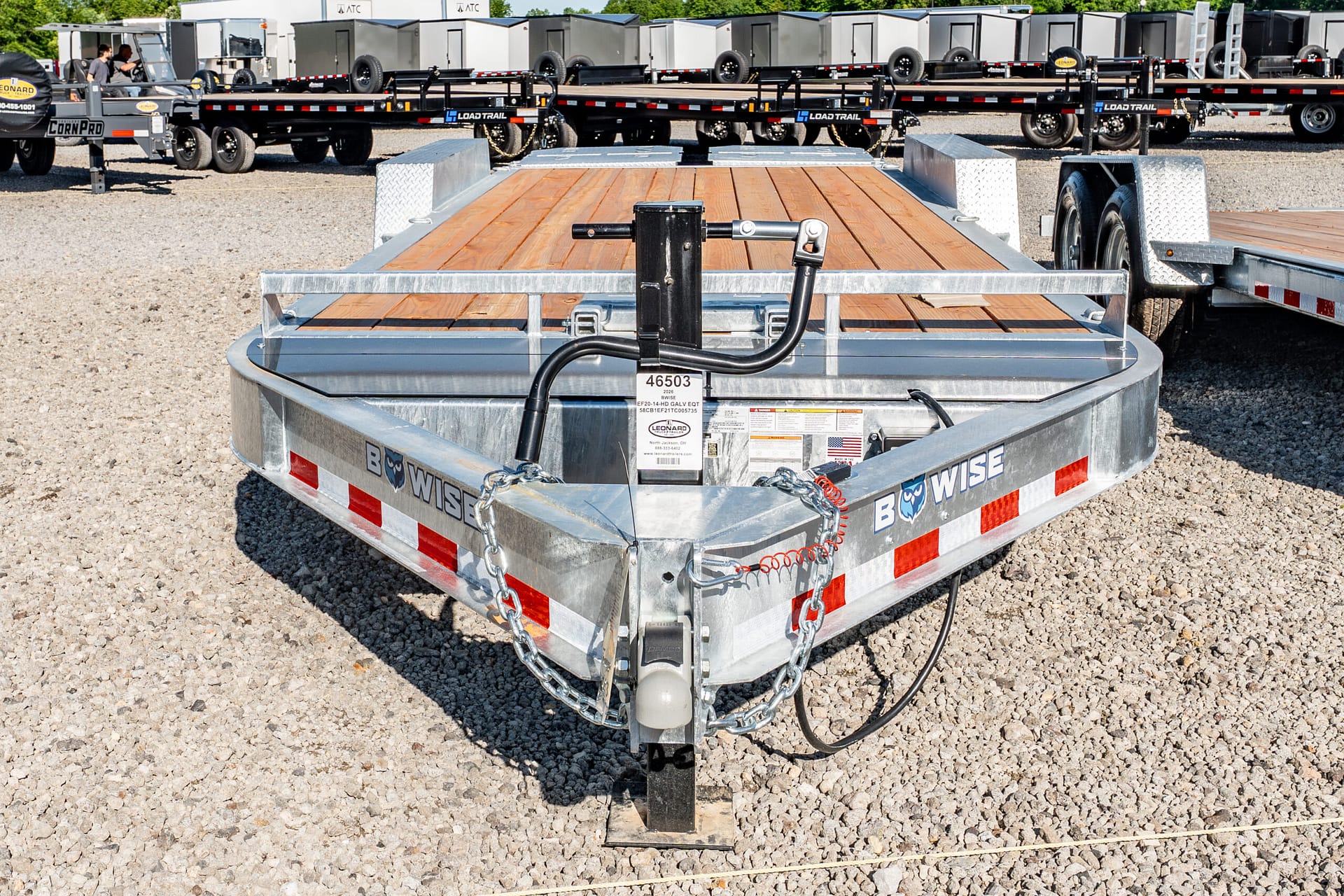 Close-up of a metal trailer hitch on a flatbed trailer with a wooden deck and chains, parked on a gravel lot with other trailers in the background.