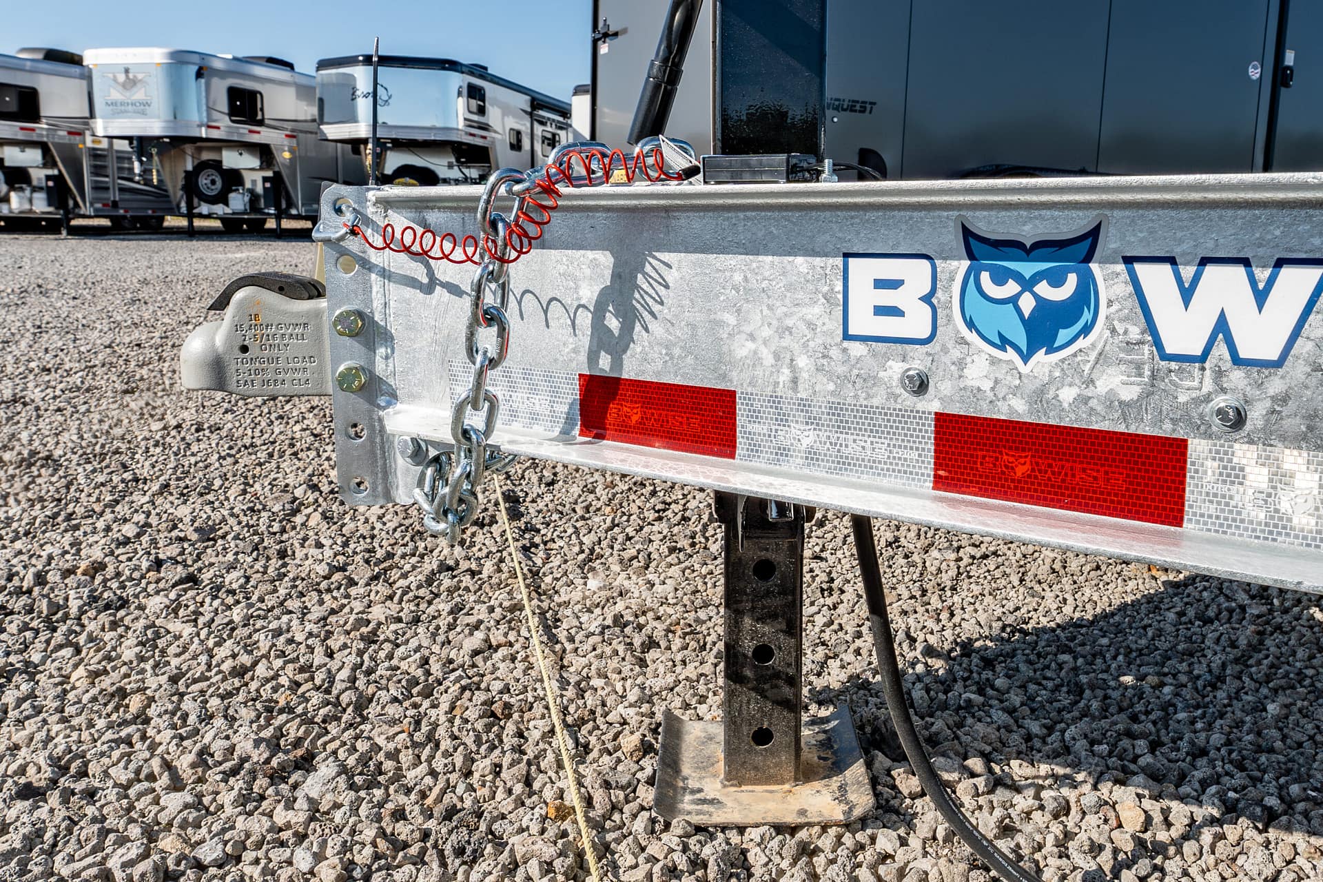 Close-up of a trailer hitch with safety chains and a red coiled cord on a gravel lot, with RV trailers in the background.