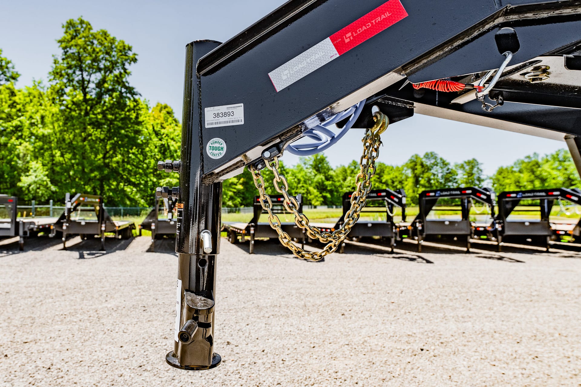 Close-up of a trailer hitch assembly with chains and a red safety sticker, set against a gravel lot and green trees in the background.