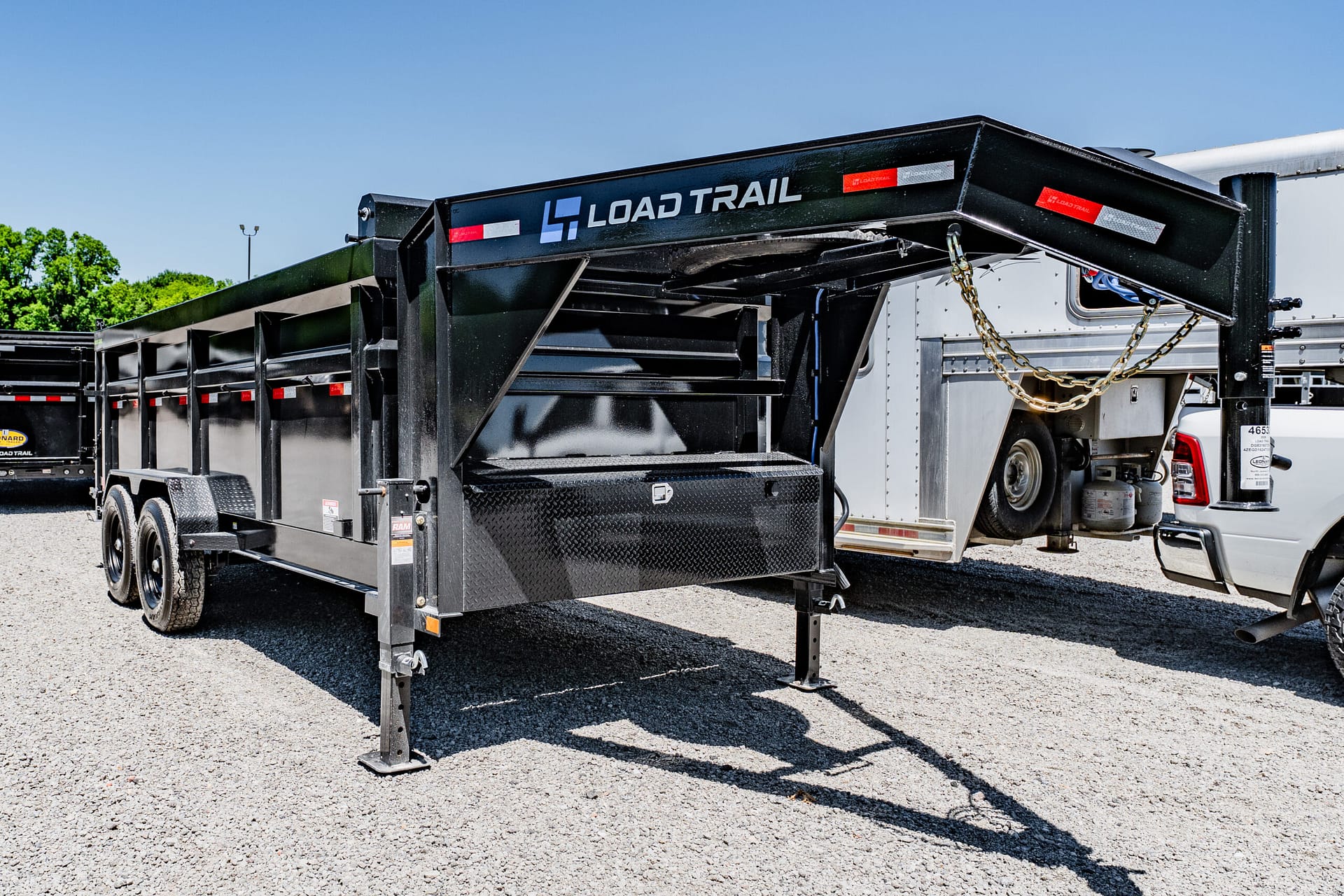Black Load Trail dump trailer with raised bed and safety chains on a gravel lot under a clear blue sky.