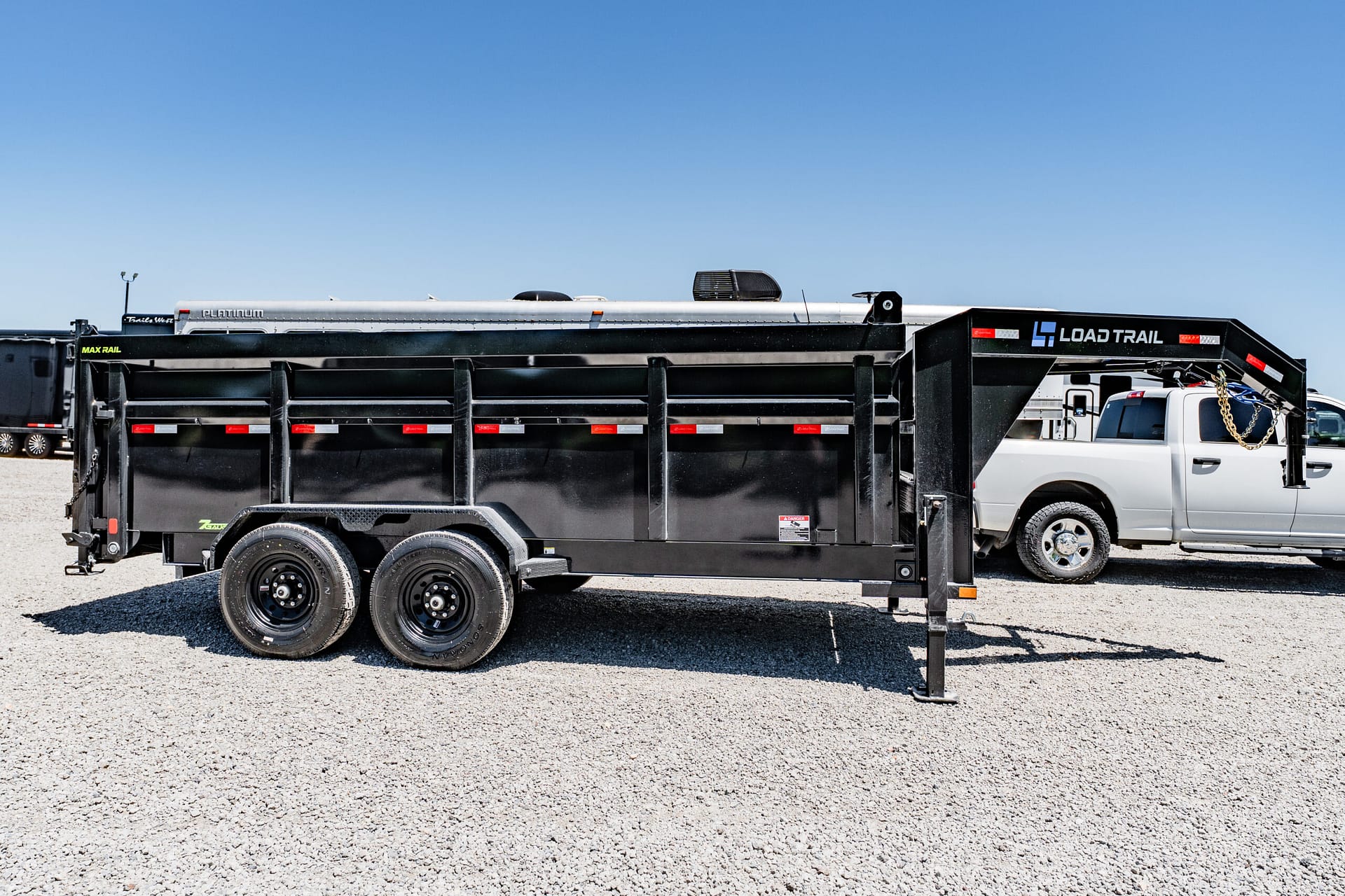 Black Load Trail dump trailer hitched to a white pickup on a gravel lot beneath a clear blue sky, showing its dual-axle wheels and support legs.