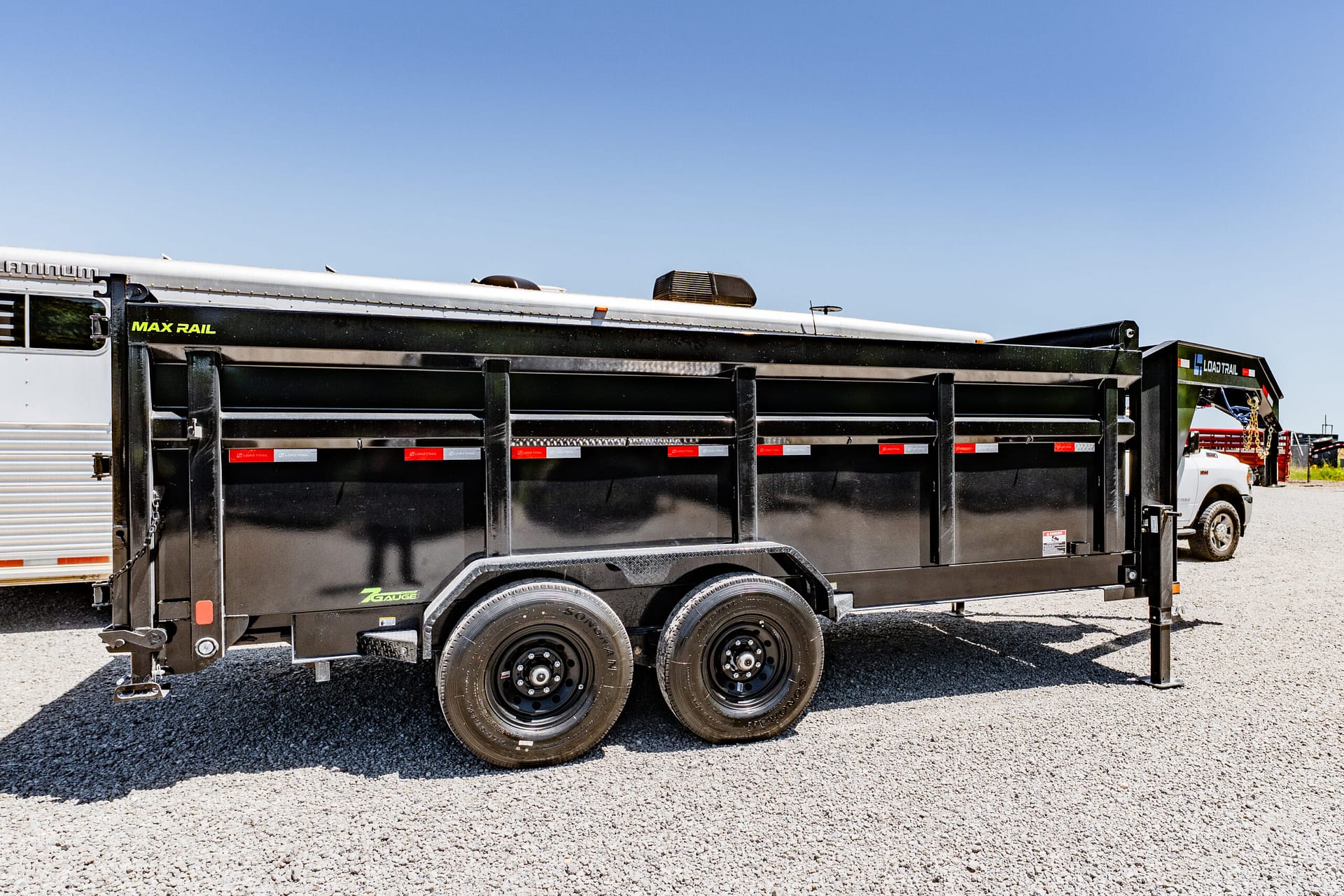 Black dump trailer with dual axles and Max Rail branding, parked on gravel under a clear blue sky.