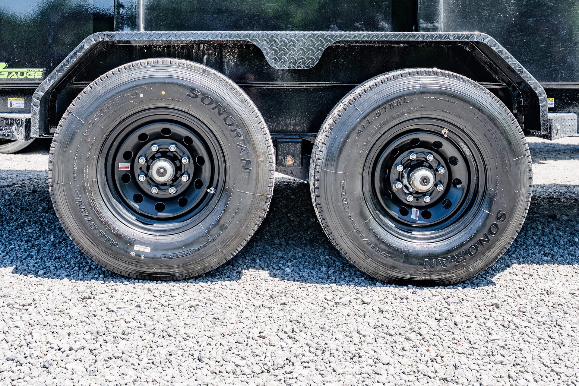 Two heavy-duty trailer wheels side by side on a black utility trailer over a gravel surface.