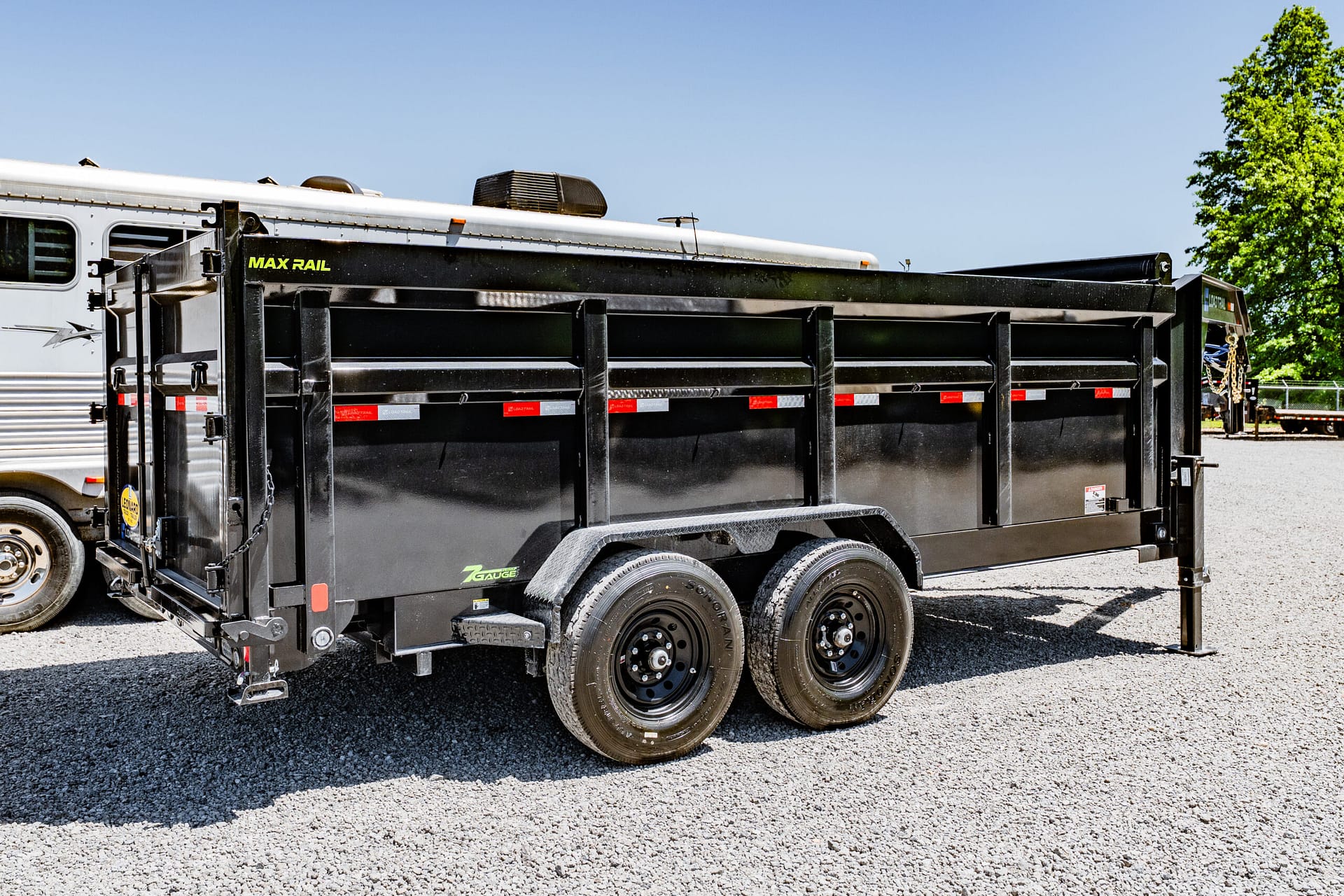 Black dump trailer labeled "MAX RAIL" hitched to a white semi-truck in a gravel lot under a clear blue sky.