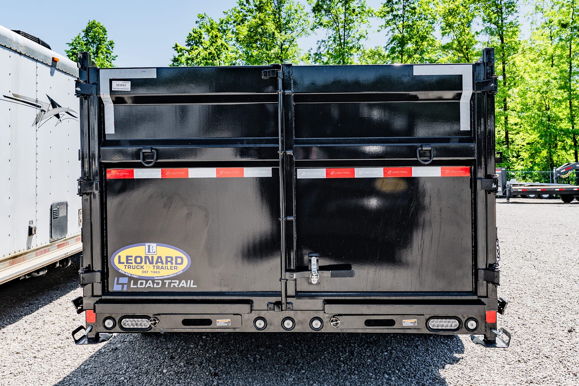 Back of a black dump trailer with red-and-white reflective tape and a Leonard Truck & Trailer sticker, set on a gravel lot with green trees in the background.