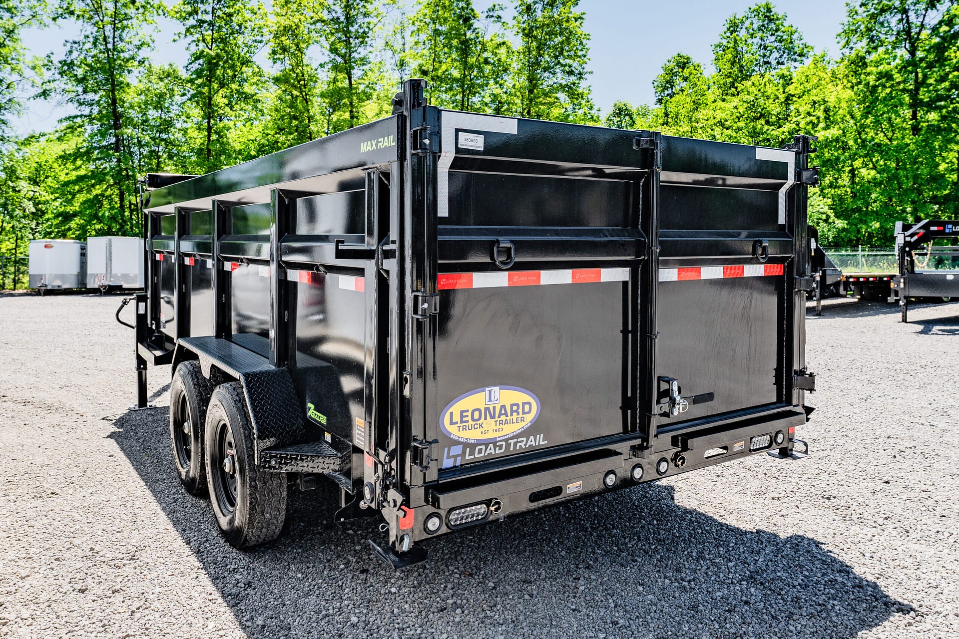 Rear view of a large black dump trailer with red-and-white safety stripes parked on a gravel lot, green trees in the background.