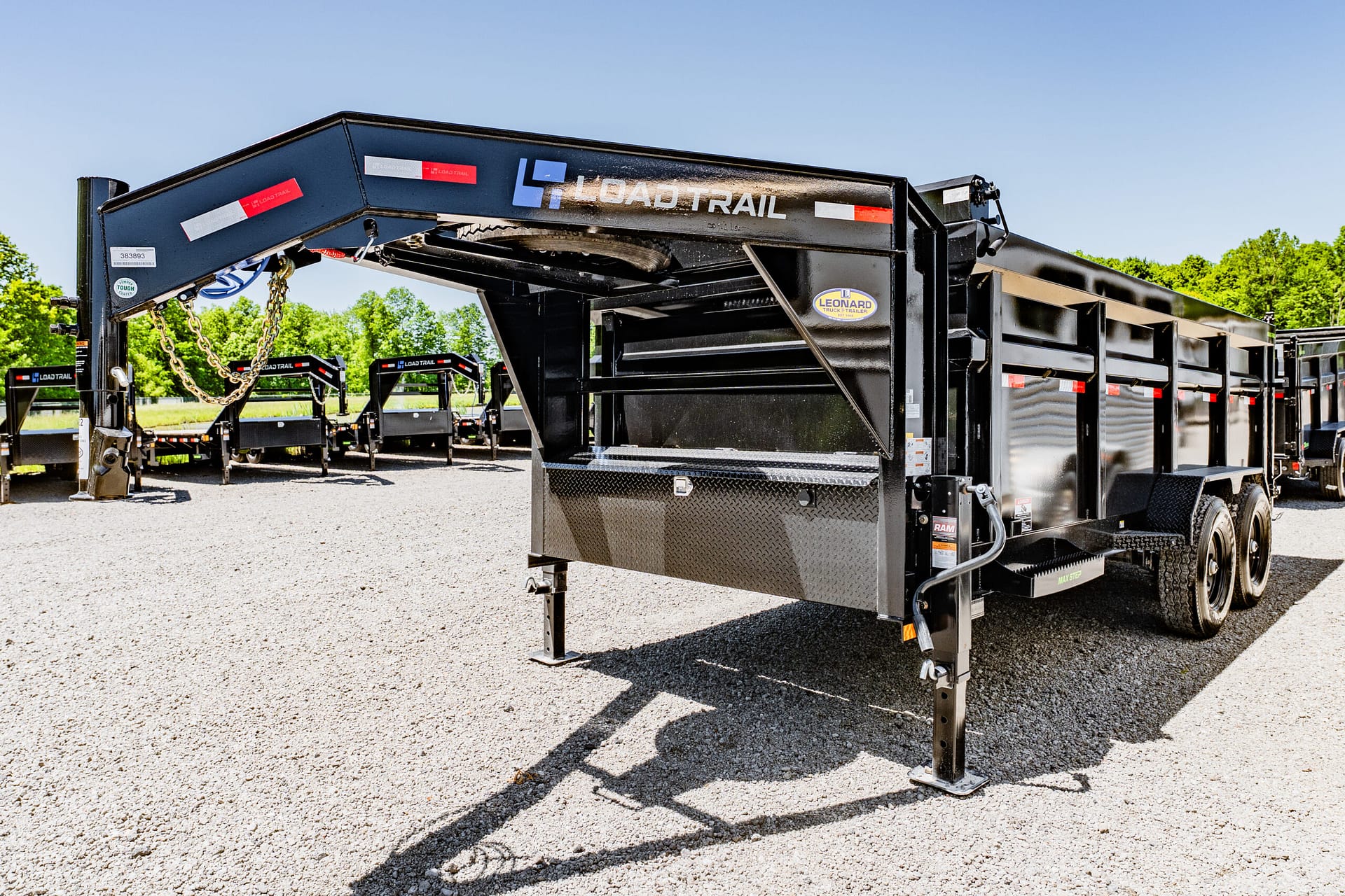 Black Load Trail heavy-duty trailer in a gravel lot, with chains and a raised front section, other trailers in the background, and a clear blue sky behind trees.