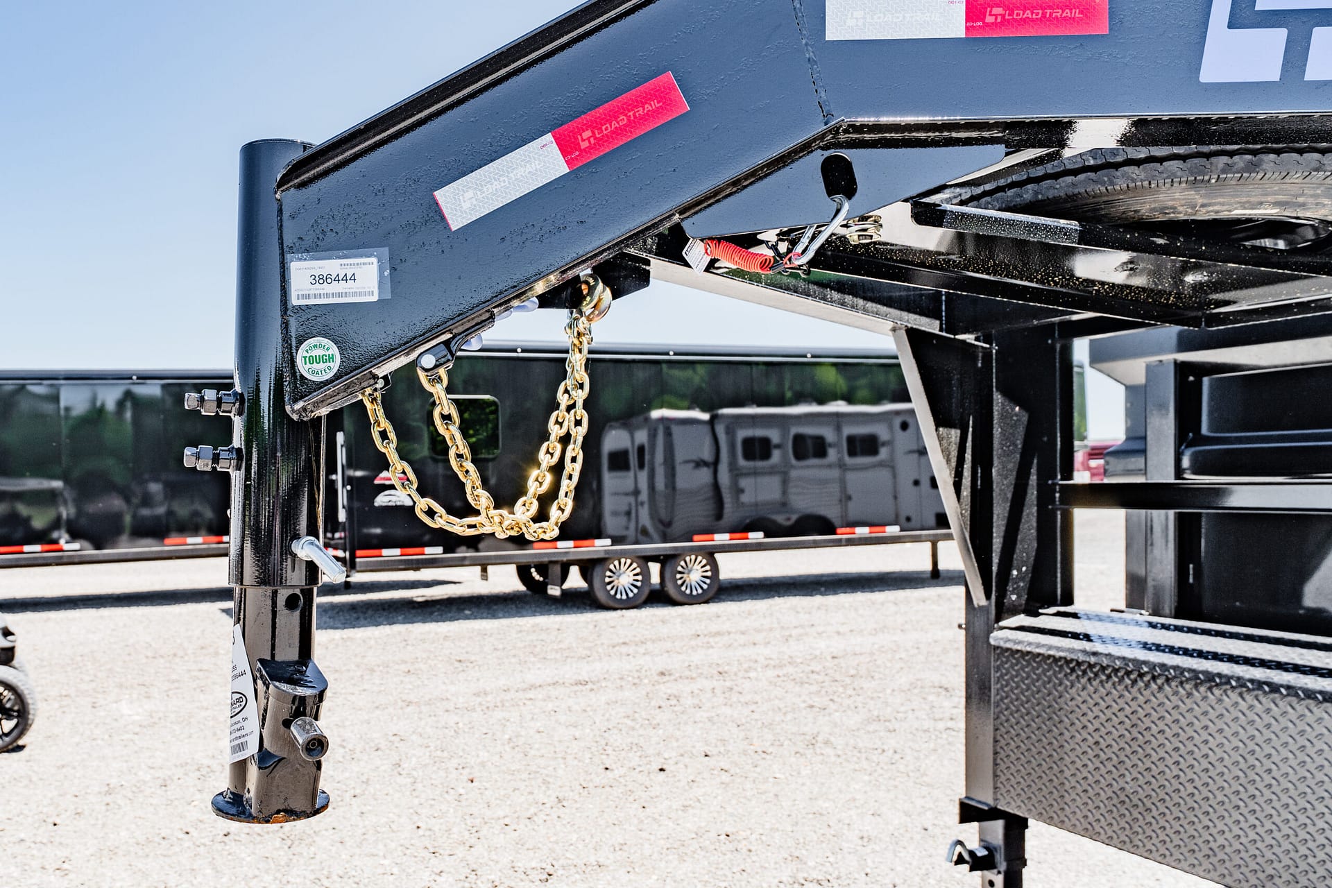 Close-up of a trailer hitch with gold safety chains and a black steel frame, parked on a gravel lot under a blue sky.