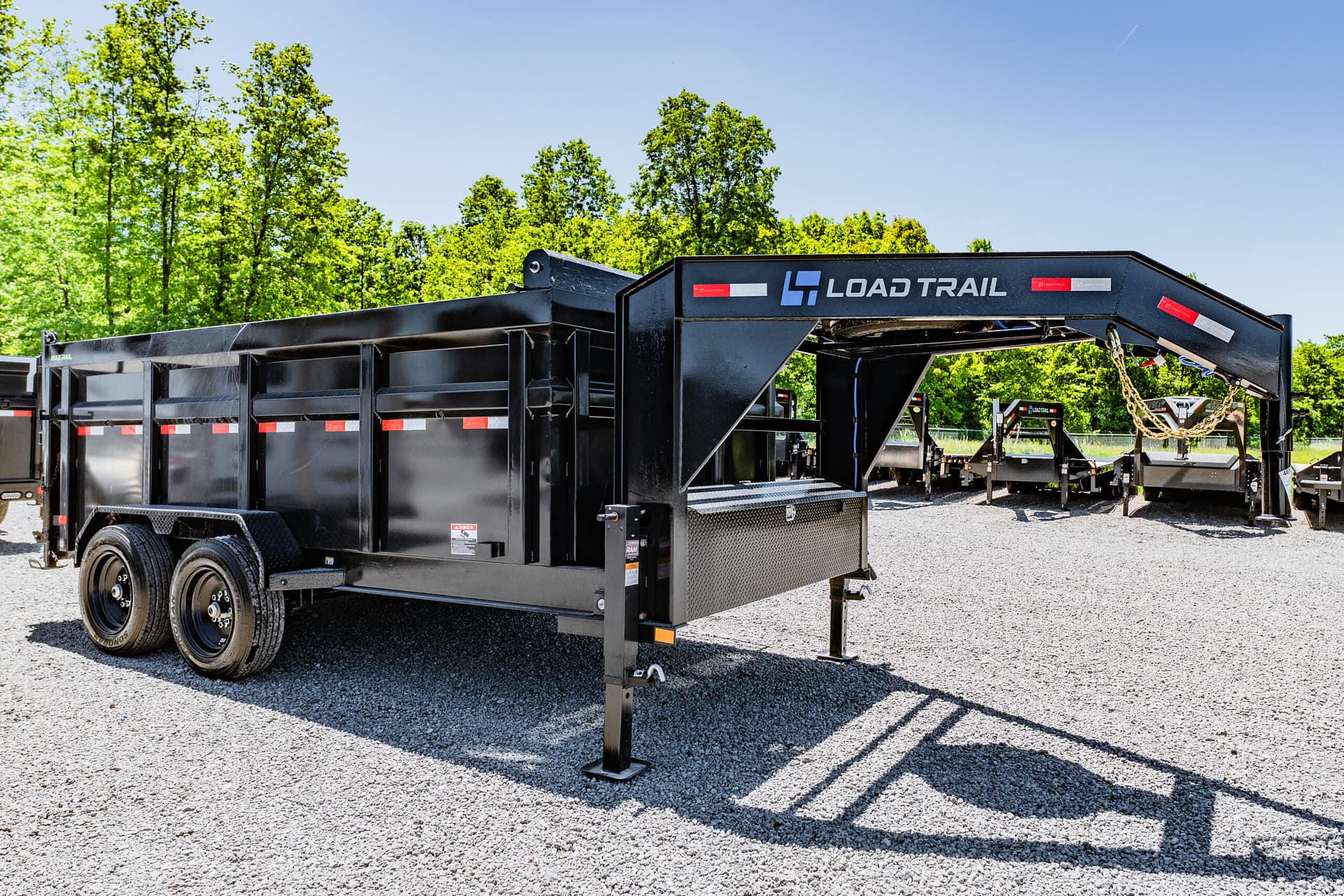 Black Load Trail dump trailer with gooseneck hitch on a gravel lot, backed by green trees under a clear blue sky.