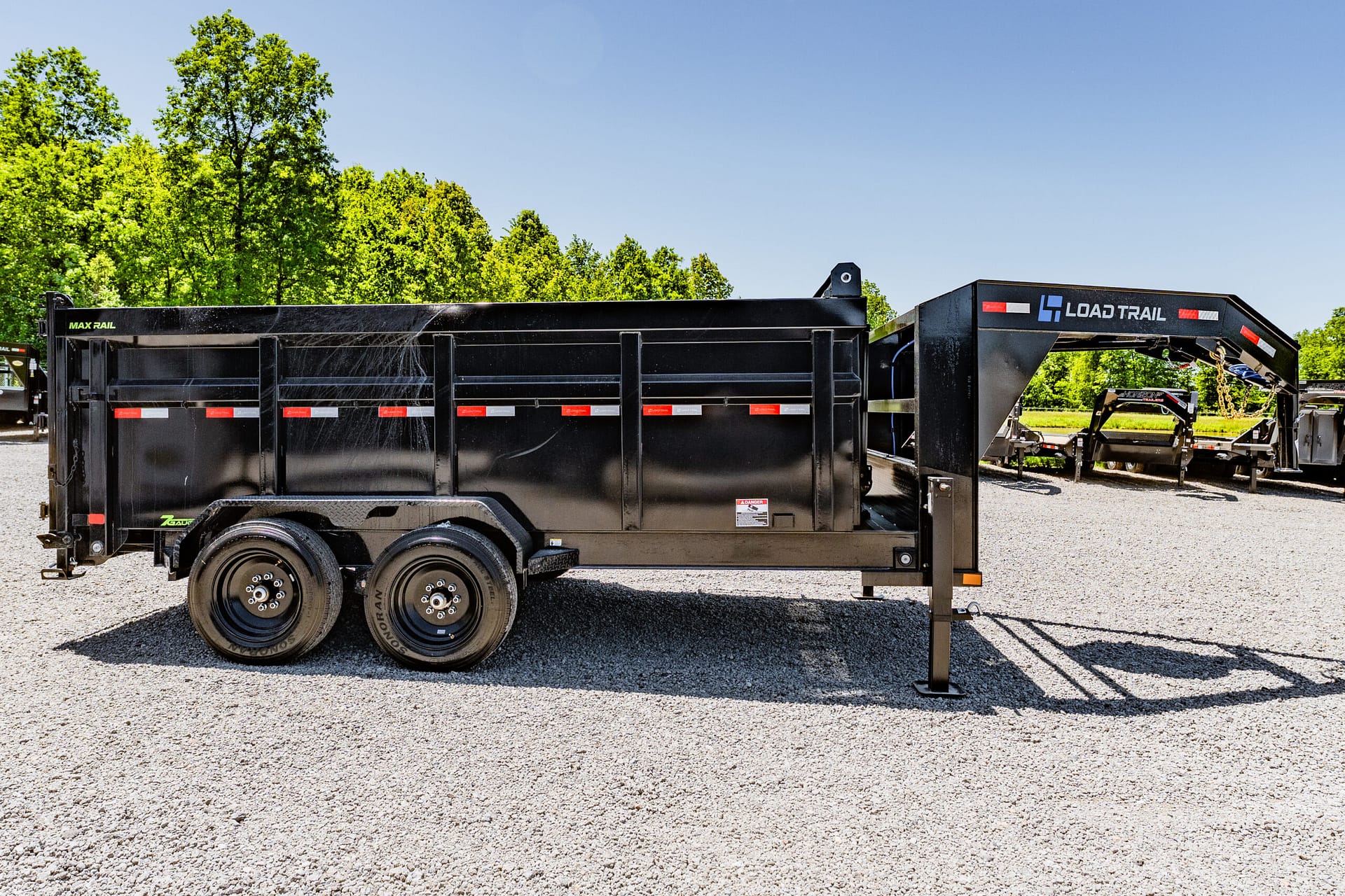 Side view of a large black dump trailer on a gravel lot, with green trees in the background and a clear blue sky.