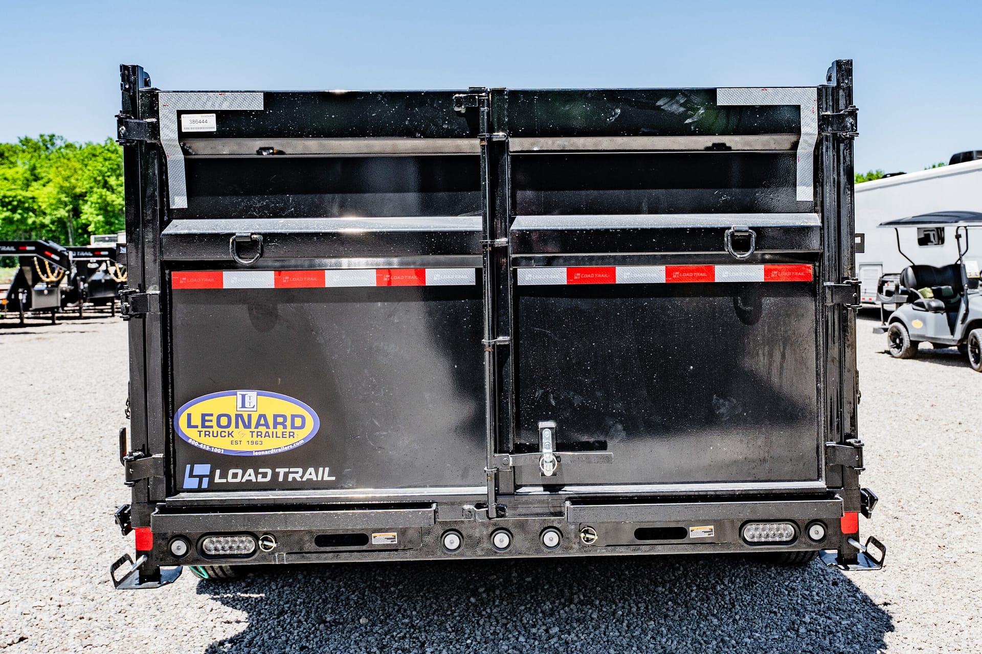 Rear view of a black Load Trail trailer with a Leonard Truck & Trailer logo, parked on a gravel lot with other trailers in the background.