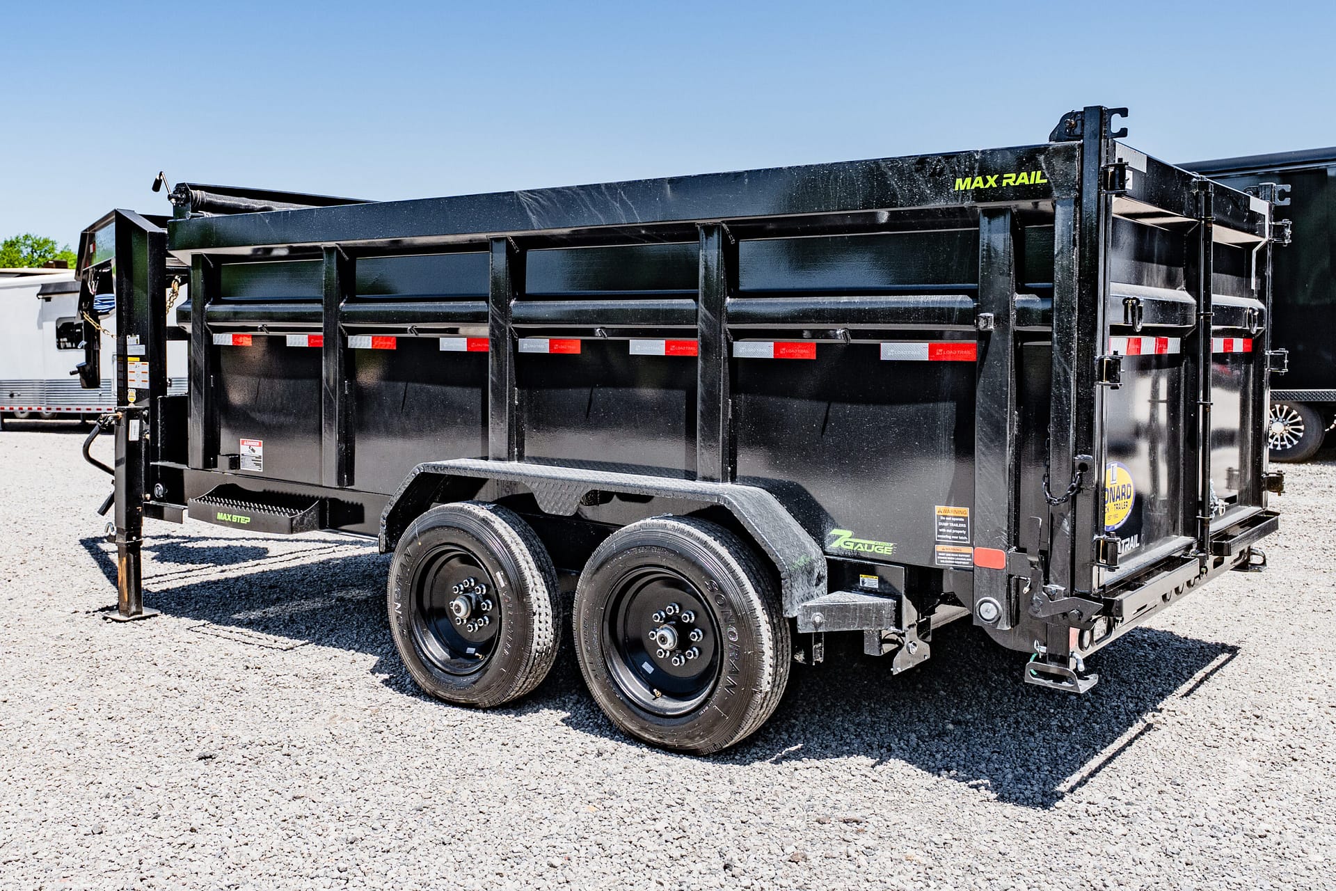 Black Max Rail dump trailer with dual axles parked on a gravel surface, showing reflective strips and raised sides.