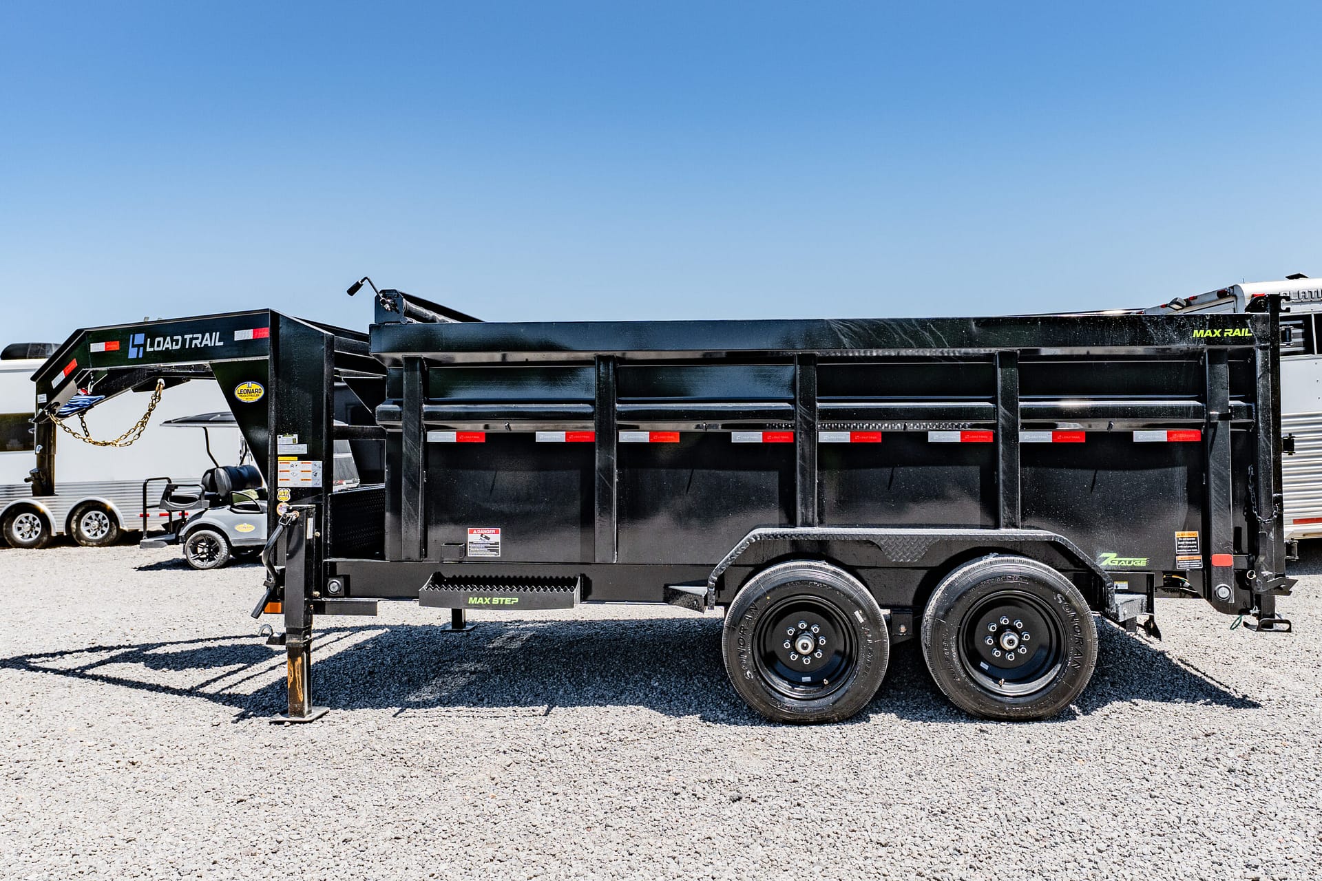 Black Load Trail dump trailer with dual axles parked on a gravel lot under a clear blue sky.