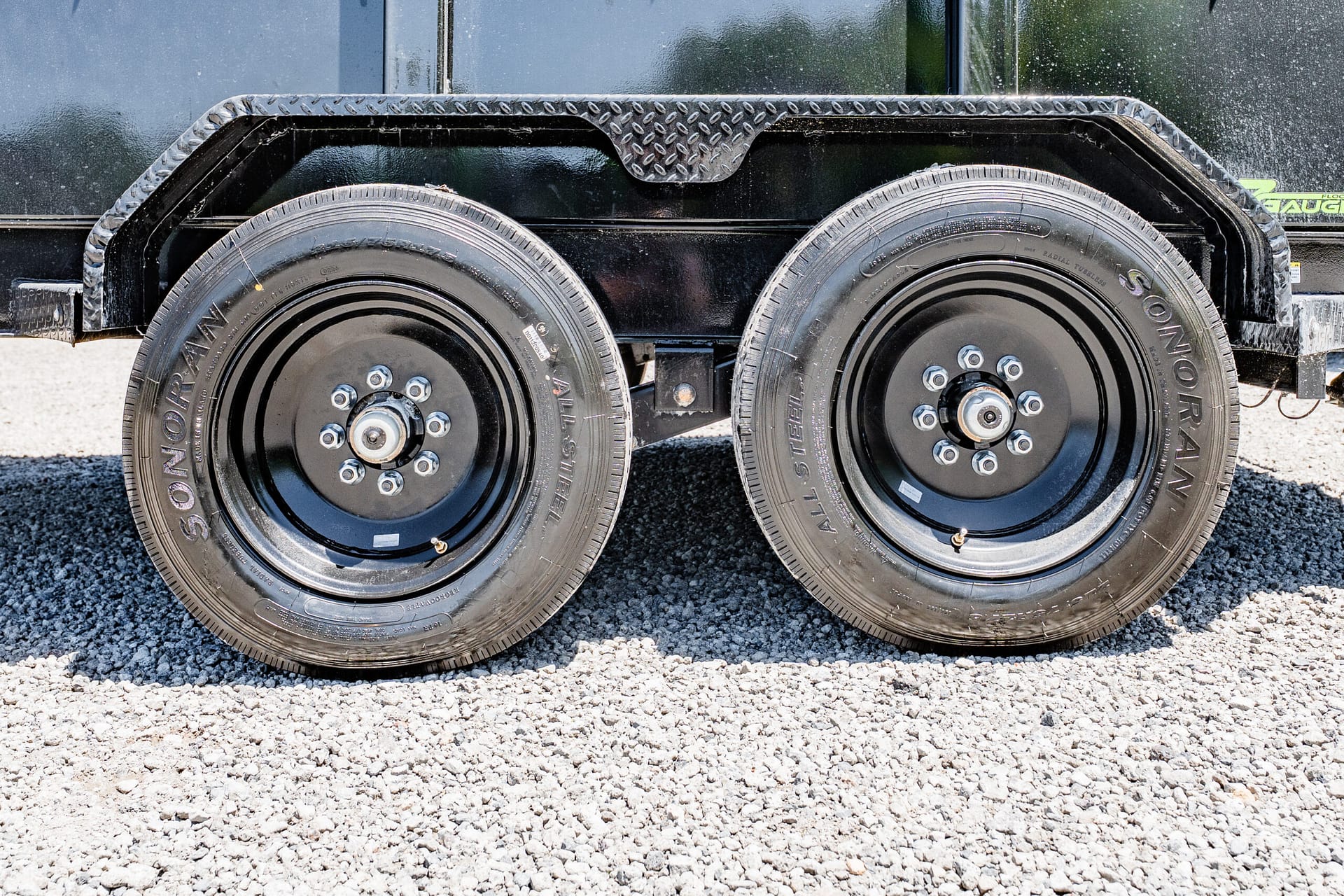 Close-up of a dual-axle trailer with black metal frame and two wheels on a gravel surface.