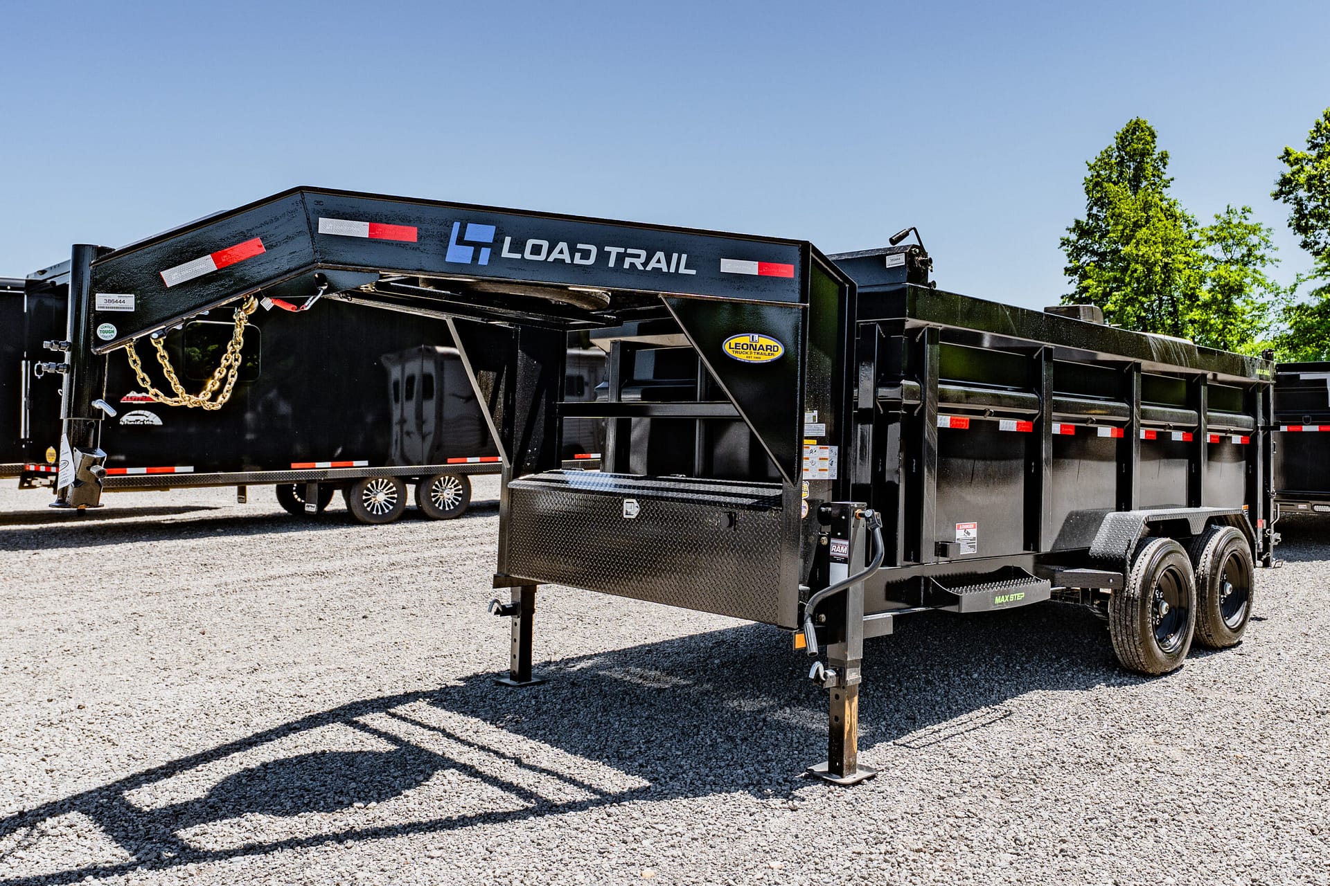 Black Load Trail dump trailer on a gravel lot, blue sky above and green trees in the background.