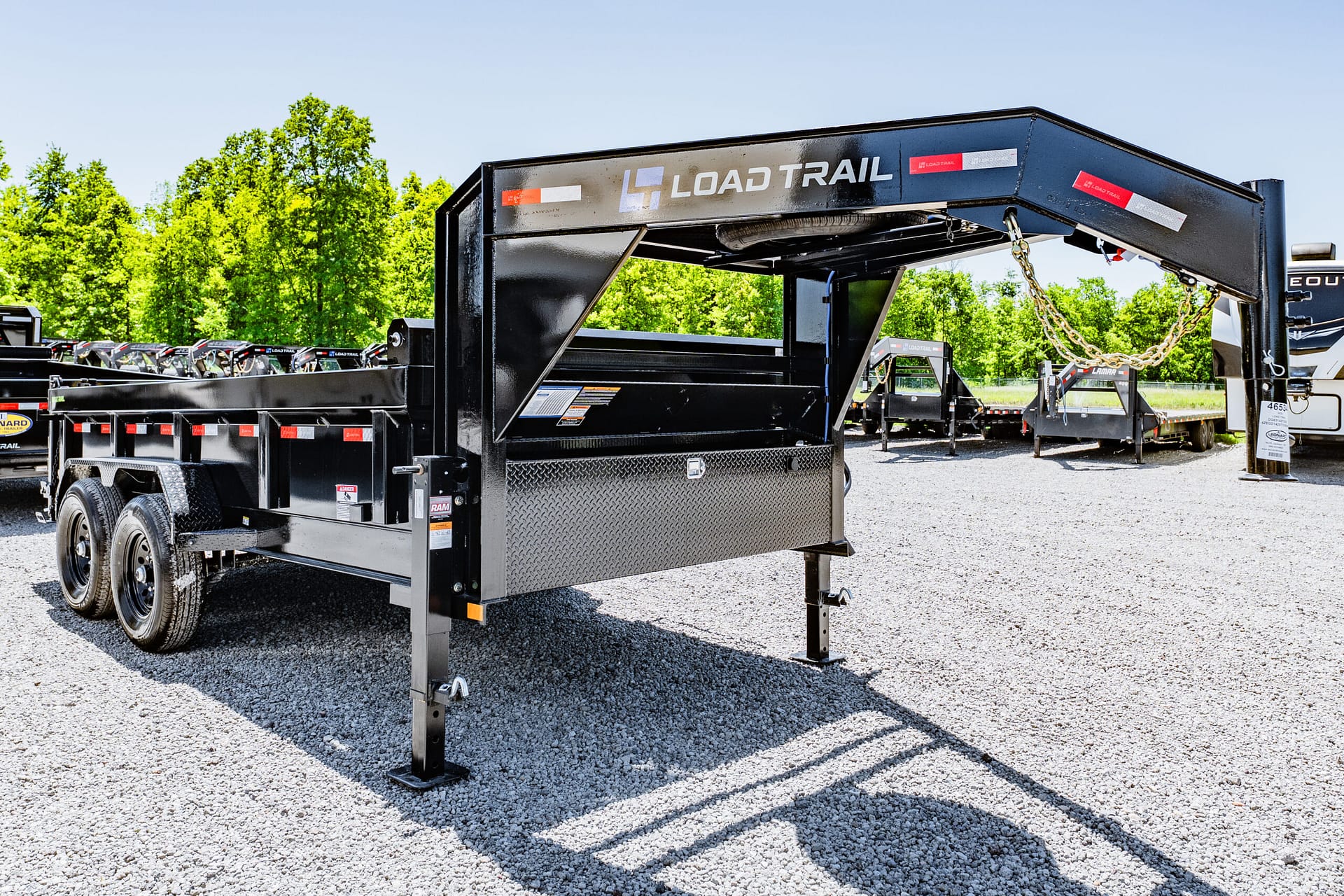 Black Load Trail dump trailer with raised bed and diamond-plate toolbox, parked on a gravel lot with green trees in the background.
