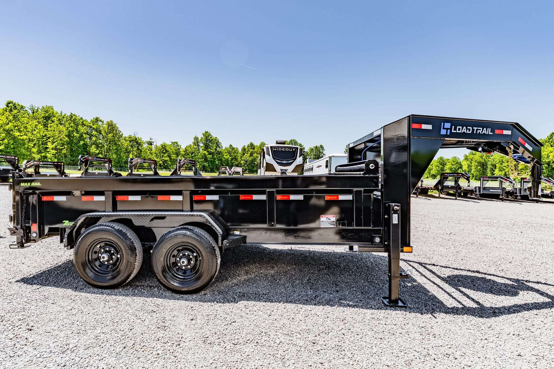 Black Load Trail utility trailer with dual axles in a gravel lot, row of trailers and a clear blue sky with green trees in the background.