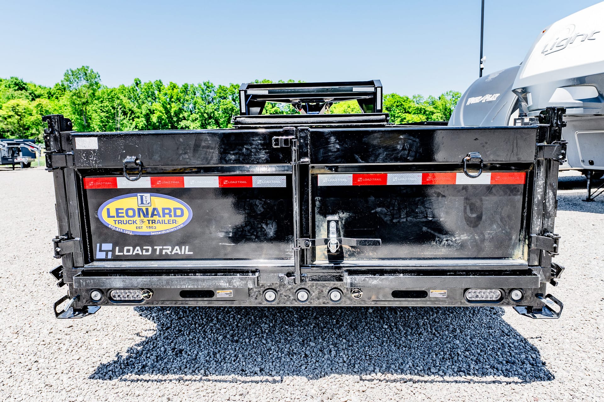Rear view of a black Load Trail dump trailer with Leonard Truck & Trailer logo, parked on a gravel lot under a clear blue sky.