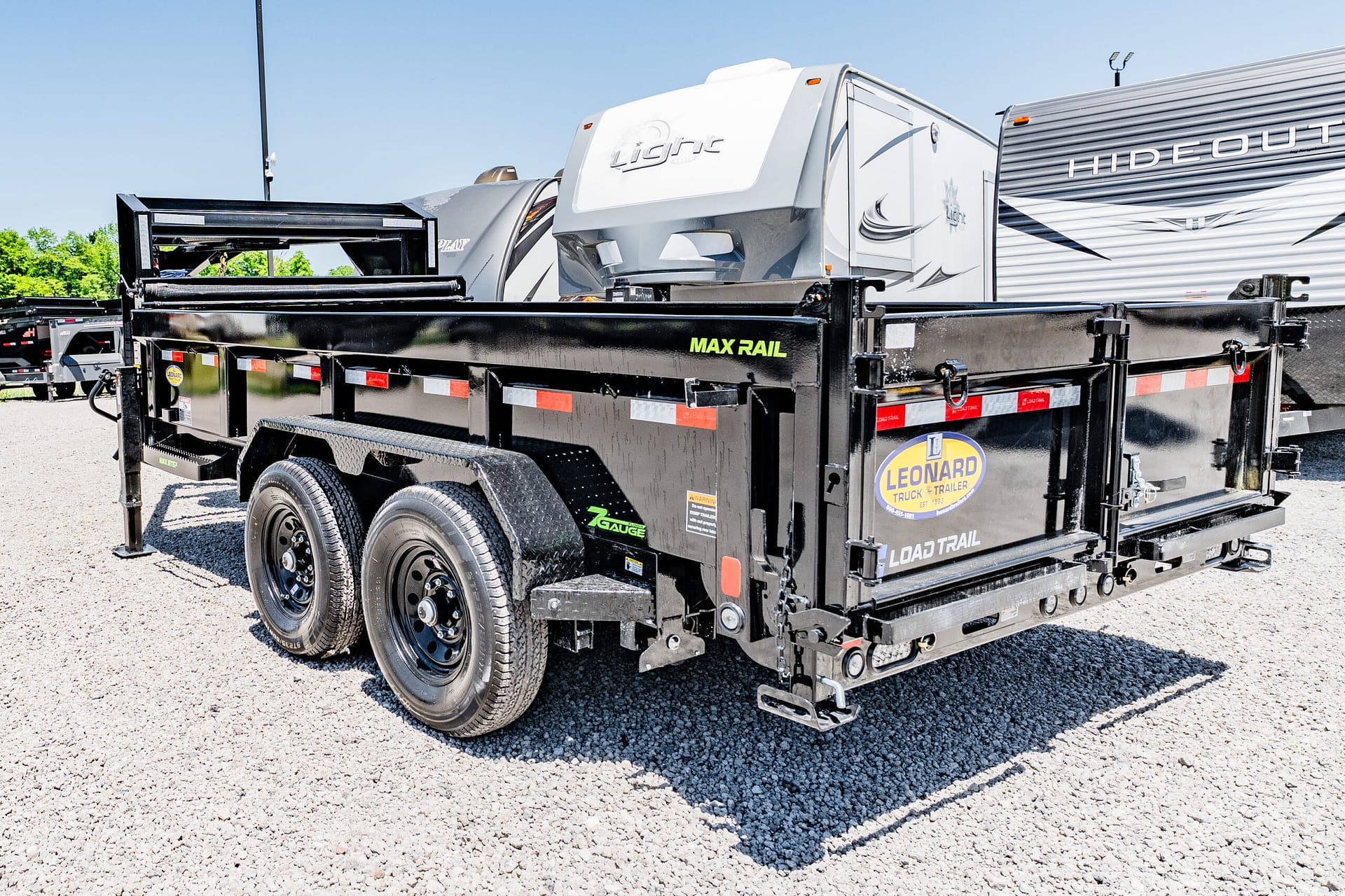 Black dual-axle trailer carrying a white travel trailer on a gravel lot, ready for transport.