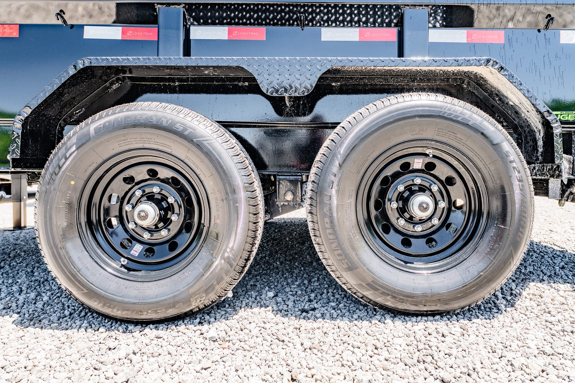 Dual trailer tires with black rims and a diamond-plate fender on a gravel lot.