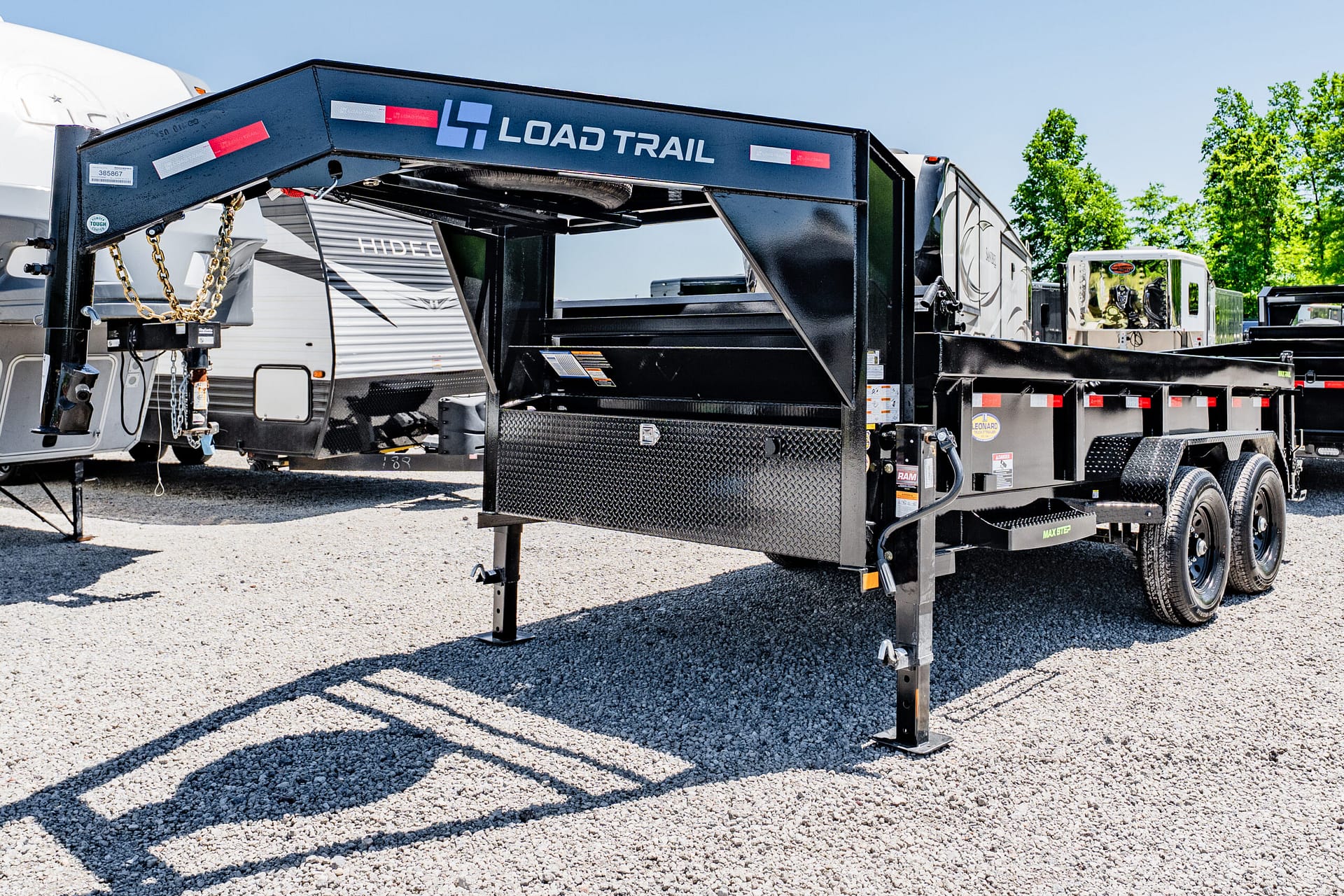 Black Load Trail gooseneck trailer frame with safety chains, toolbox, and diamond-plate steps on a gravel lot.