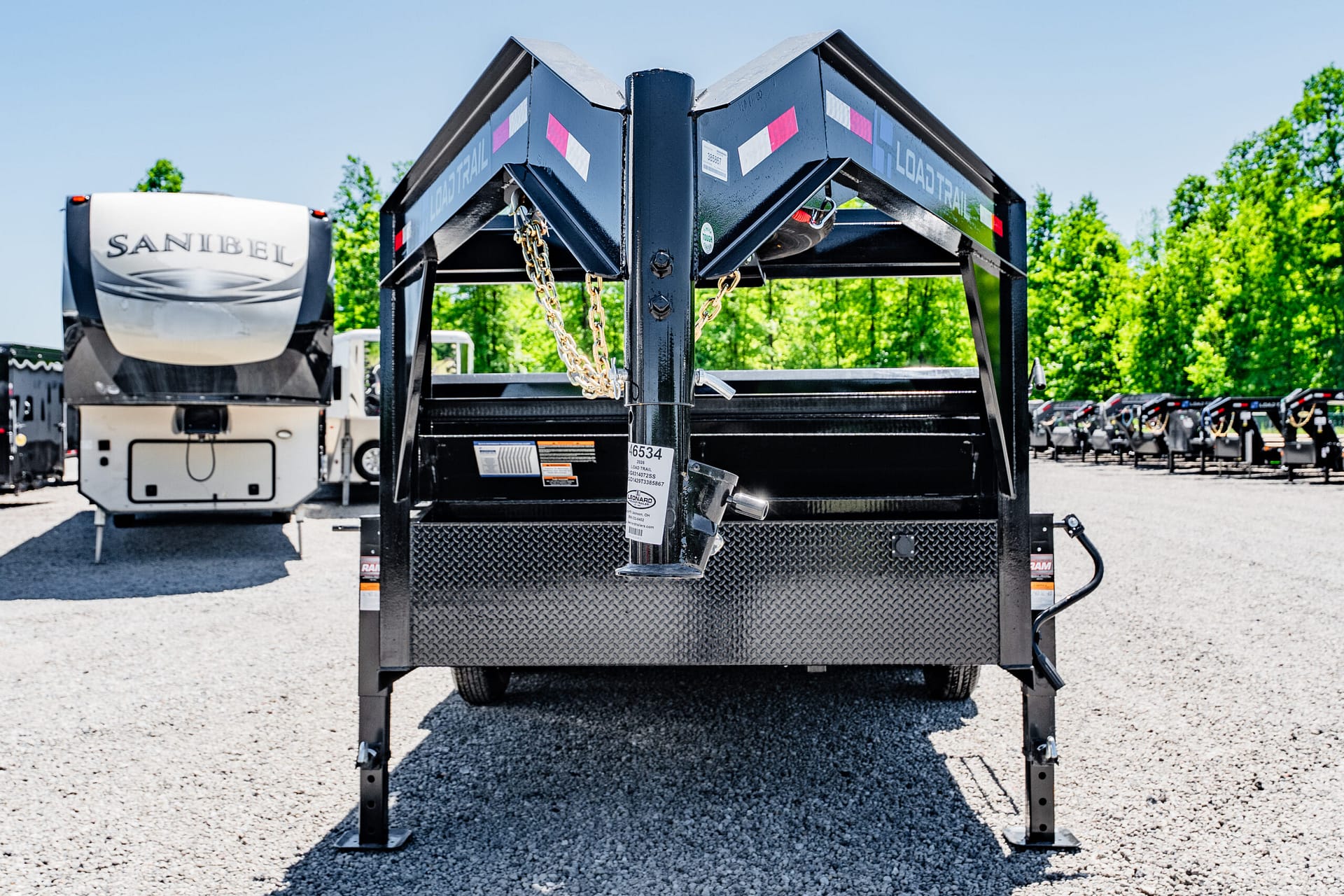 Front view of a black load-trail trailer tongue with safety chains, in a dealership lot with gravel ground and green trees in the background
