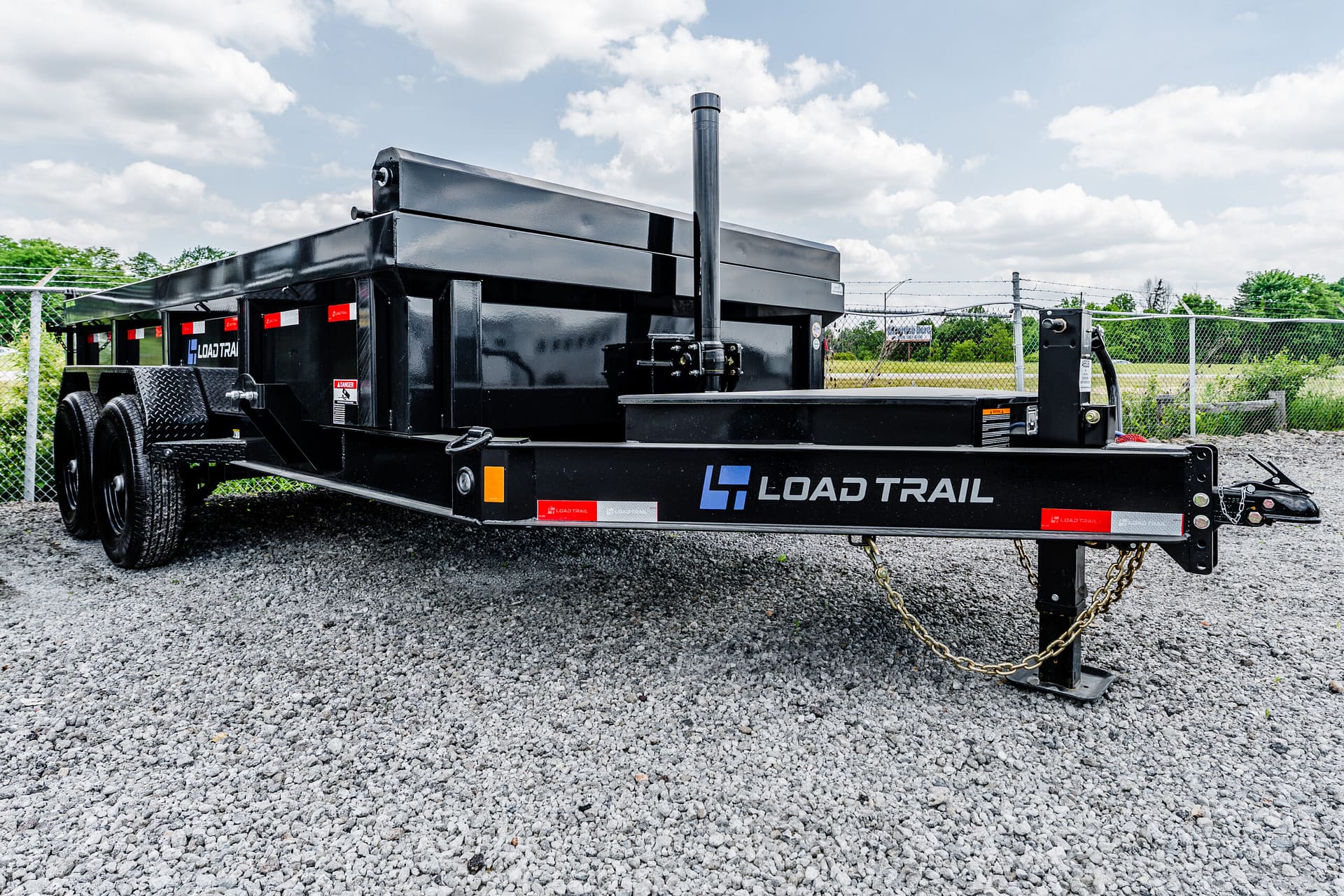 Black Load Trail dump-style utility trailer on a gravel lot with a chain hitch and chain barrier in the foreground, fence and trees in the background, under a partly cloudy sky.