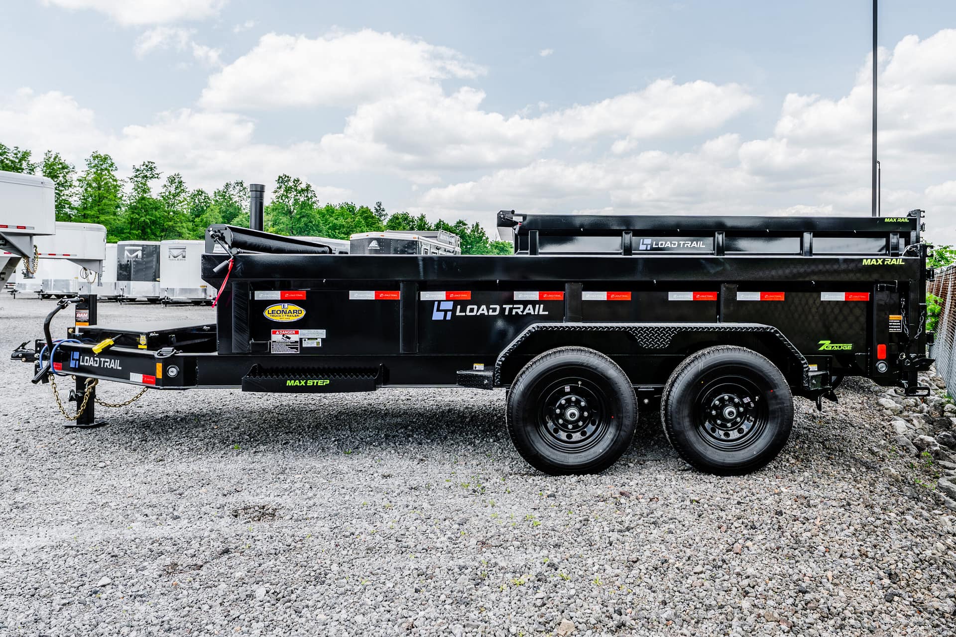 Black Load Trail utility trailer with dual axles on a gravel lot, logo visible and white trailers in the background.