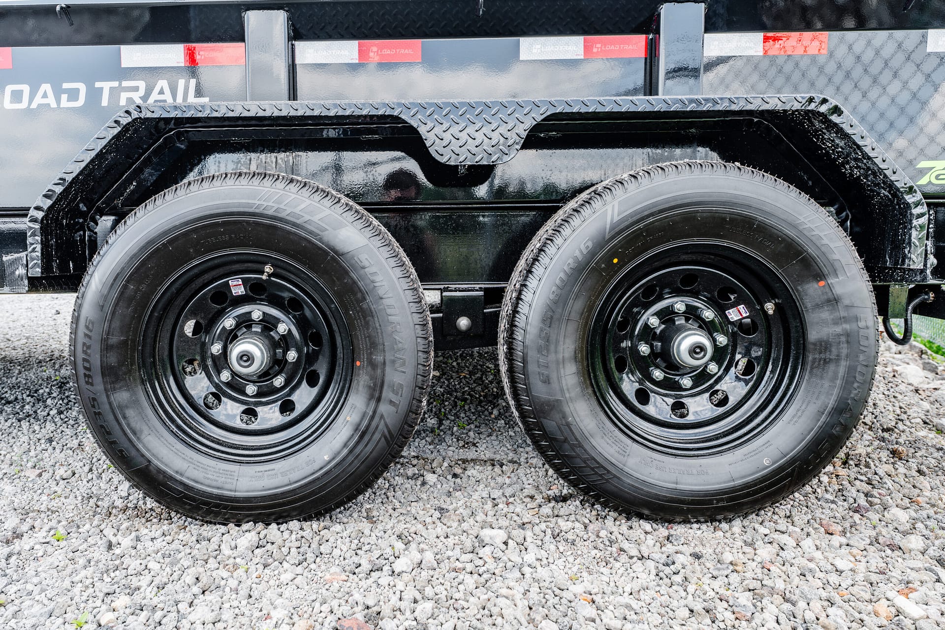 Dual-axle trailer with large black tires and diamond-plate fenders on gravel surface.