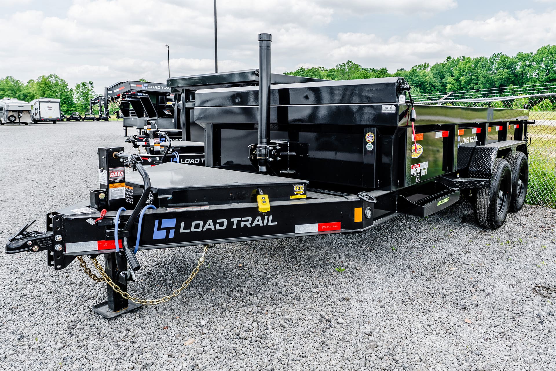 Black Load Trail heavy-duty trailer on a gravel lot, with a chain and hitch in the foreground and other trailers in the background.