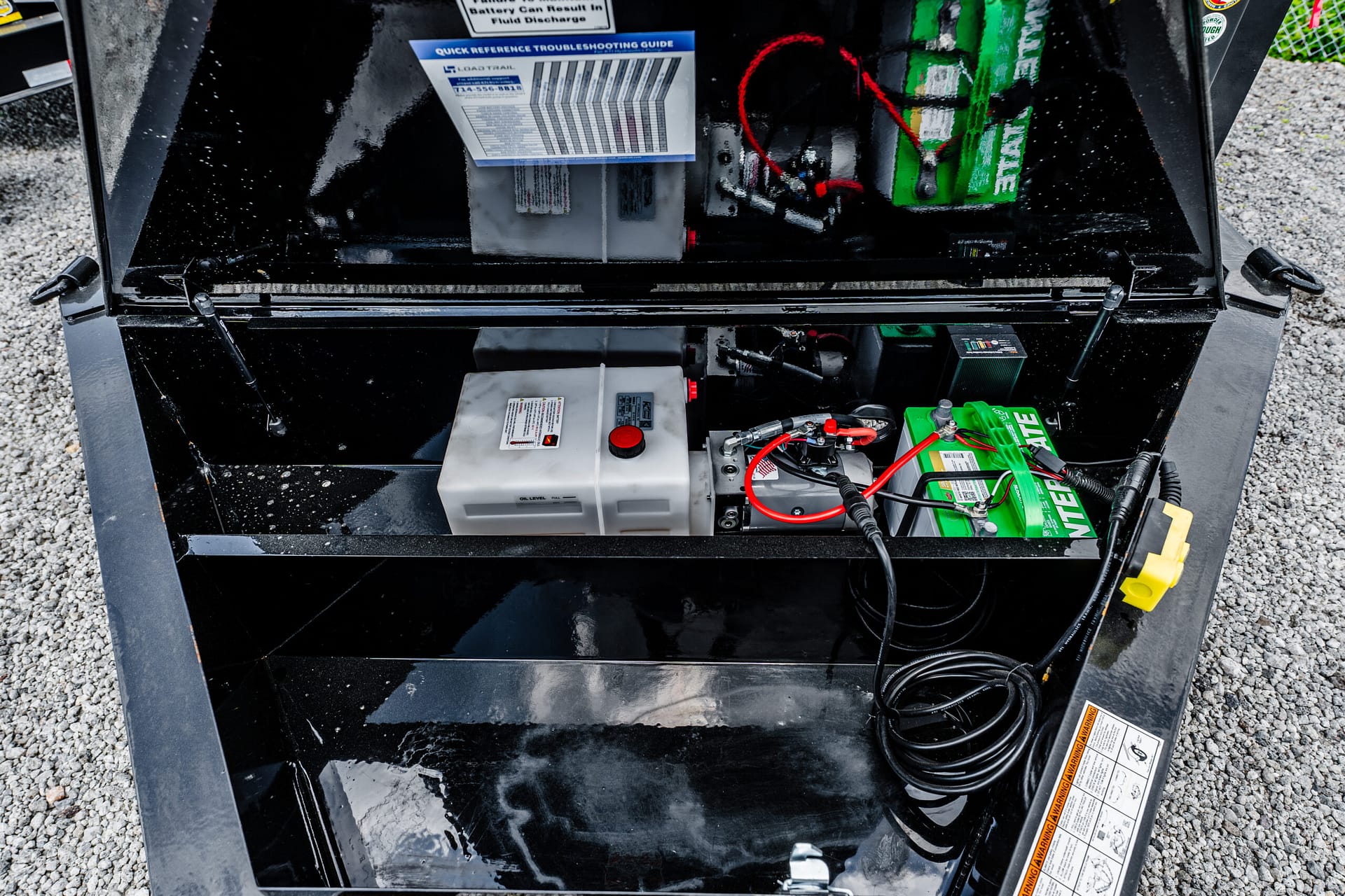 Open battery compartment with a green battery and tangled red/black cables inside a black metal box on gravel.