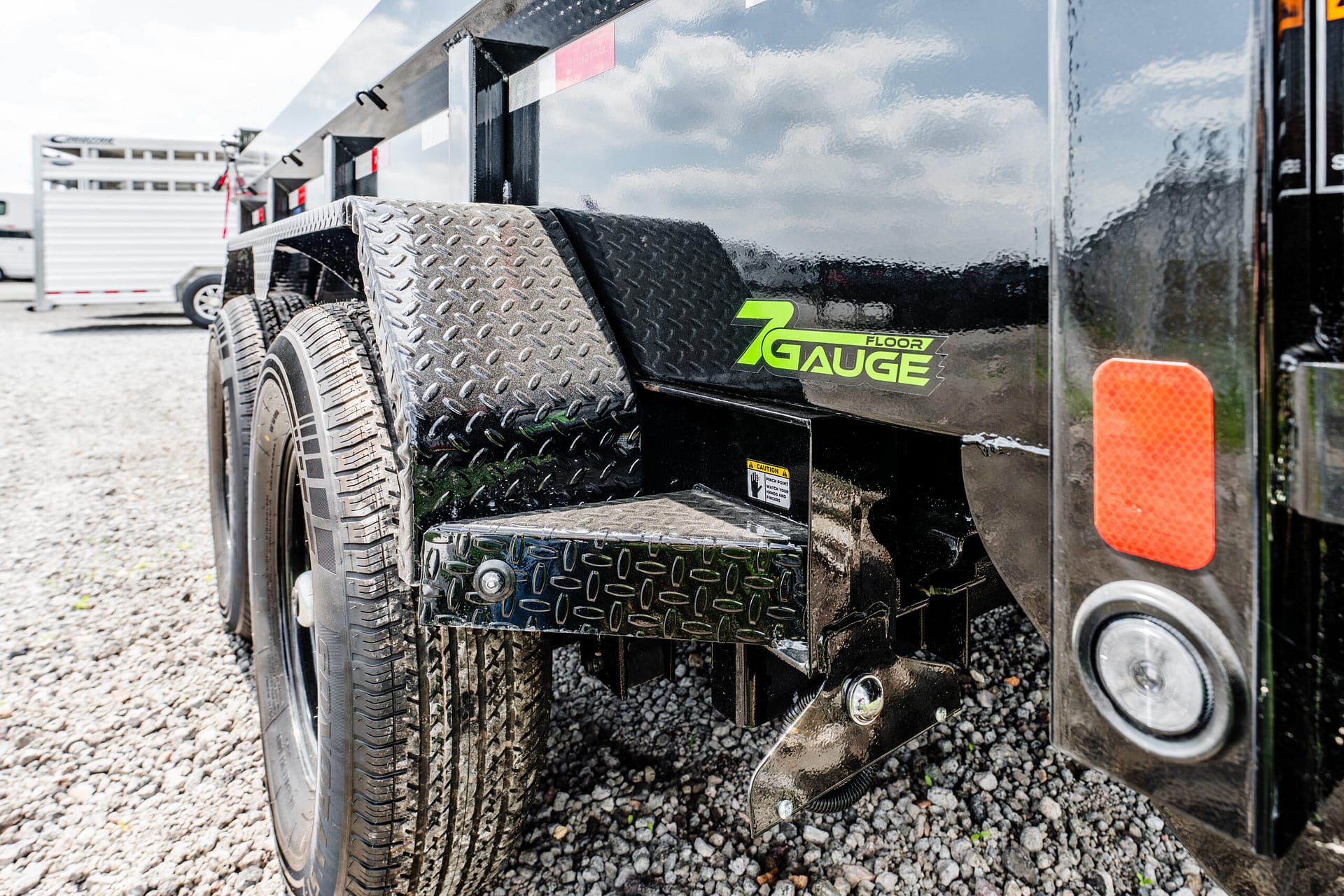 Close-up of a black trailer with a diamond-plate step, tire tread, and a bright green '7 GAUGE FLOOR' badge on the side guard.
