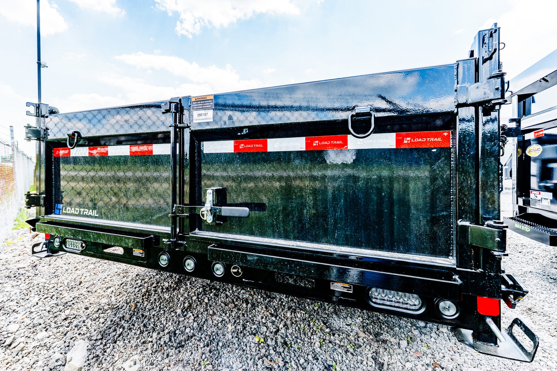 Rear view of a black Load Trail dump trailer on gravel, showing red/white reflective stripes, latches, and safety rings.