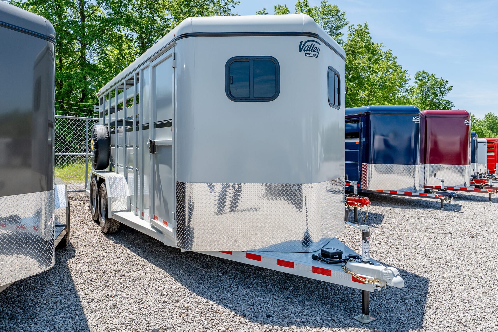 Row of horse trailers parked on a gravel lot, with a light gray trailer in the foreground and green trees in the background.