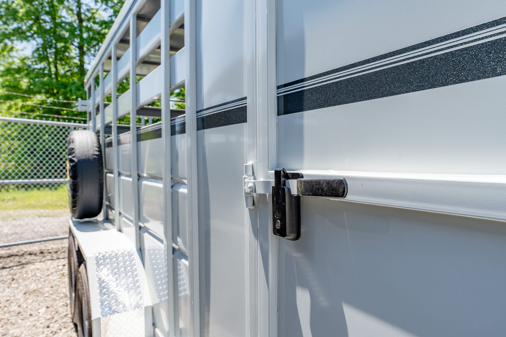 Side view of a light gray trailer with a black glitter stripe, ladder-like vents, and a mounted latch; spare tire and chain-link fence in background.