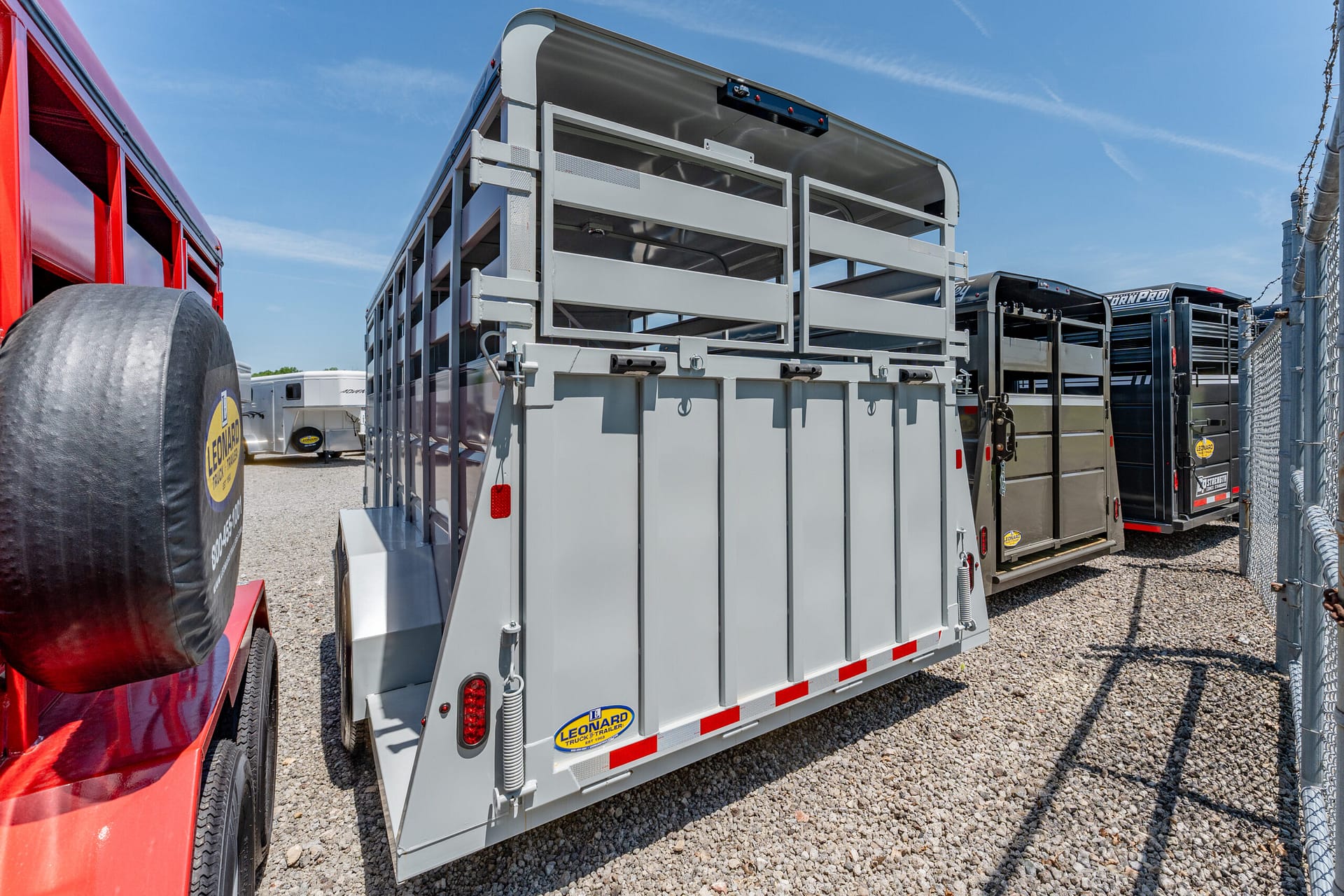 Row of grey livestock trailers parked in a gravel lot beside a chain-link fence on a sunny day.
