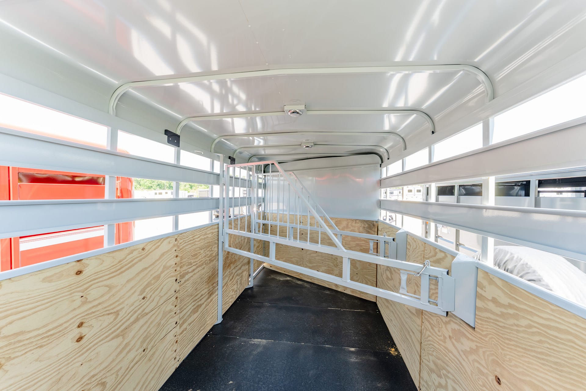 Interior of a livestock trailer: light gray metal stalls, plywood side walls, and a dark non-slip floor beneath daylight from side windows.